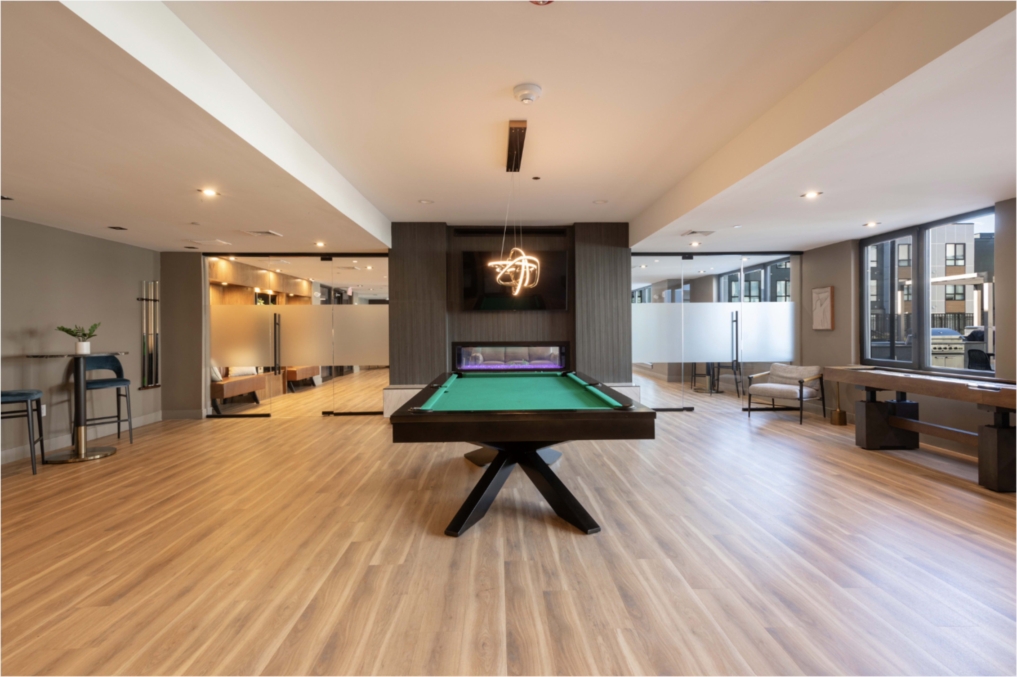 The image shows a spacious room with a green pool table centered, modern lighting fixtures, and large windows allowing natural light to illuminate the wooden floor.