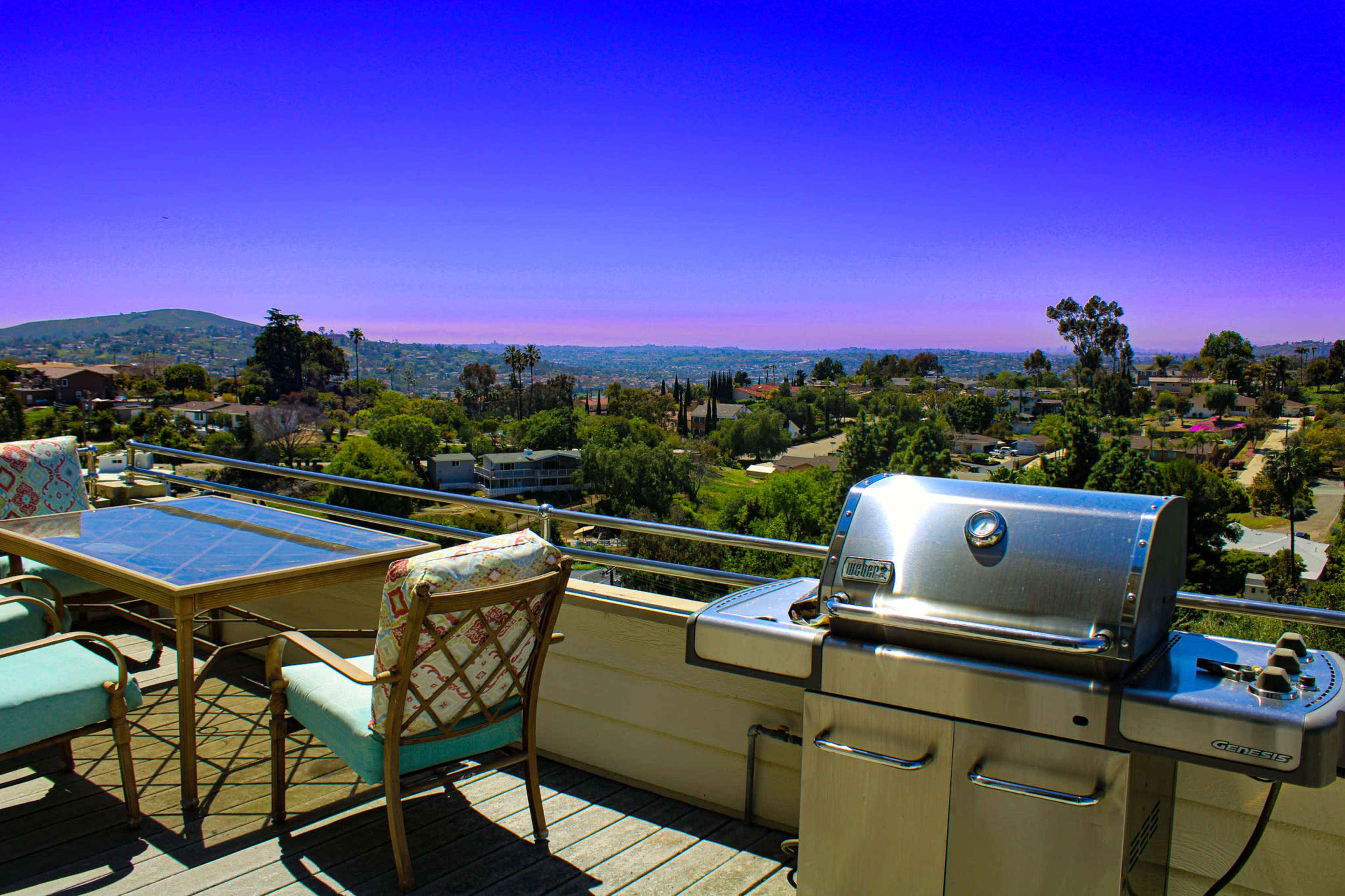 A balcony features a grill and a dining table with chairs overlooking a scenic view of the landscape and hills.