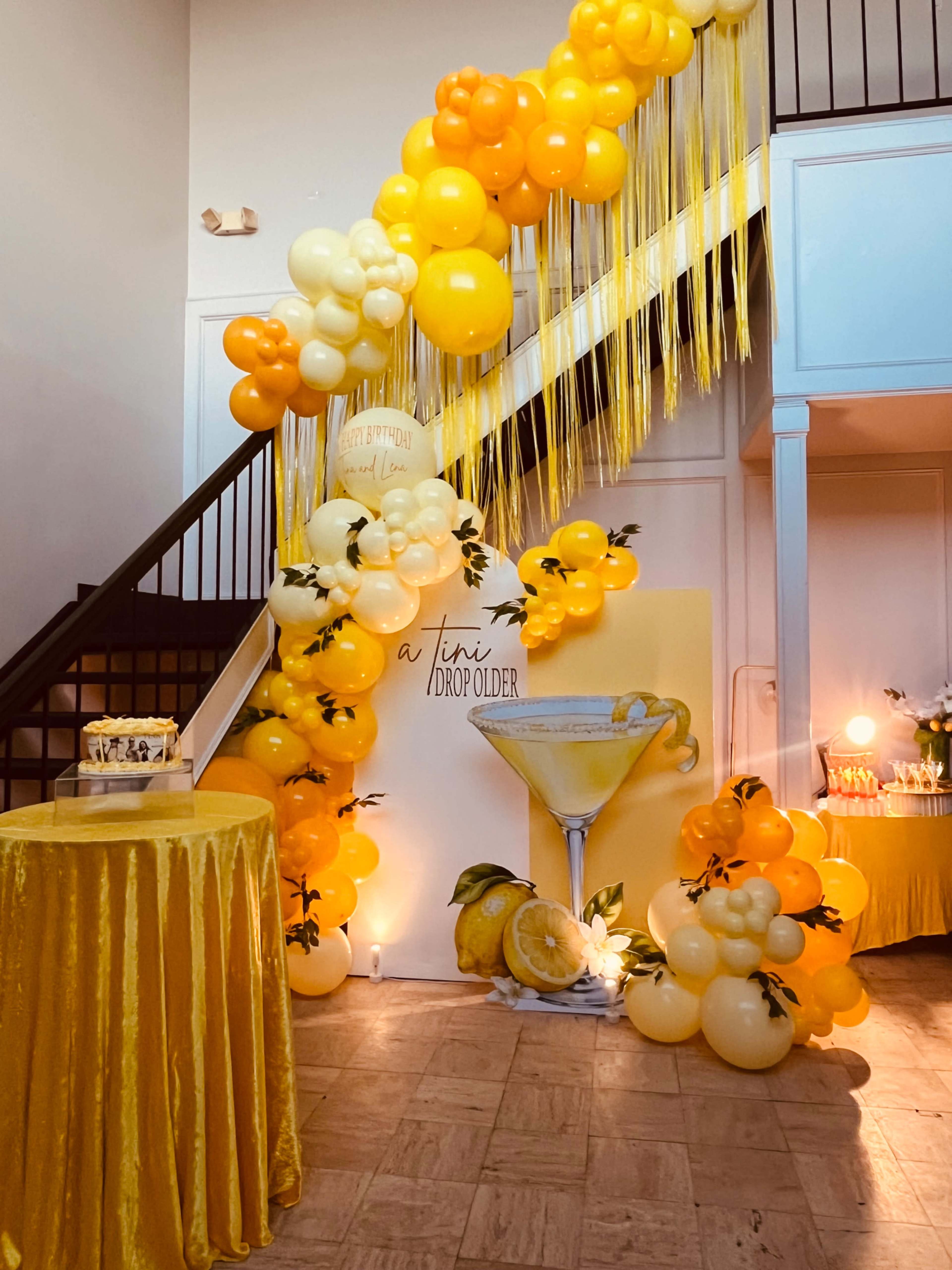 The image shows a festive birthday decoration featuring a staircase adorned with yellow and orange balloons, a backdrop with a cocktail graphic, and a table draped in yellow fabric.
