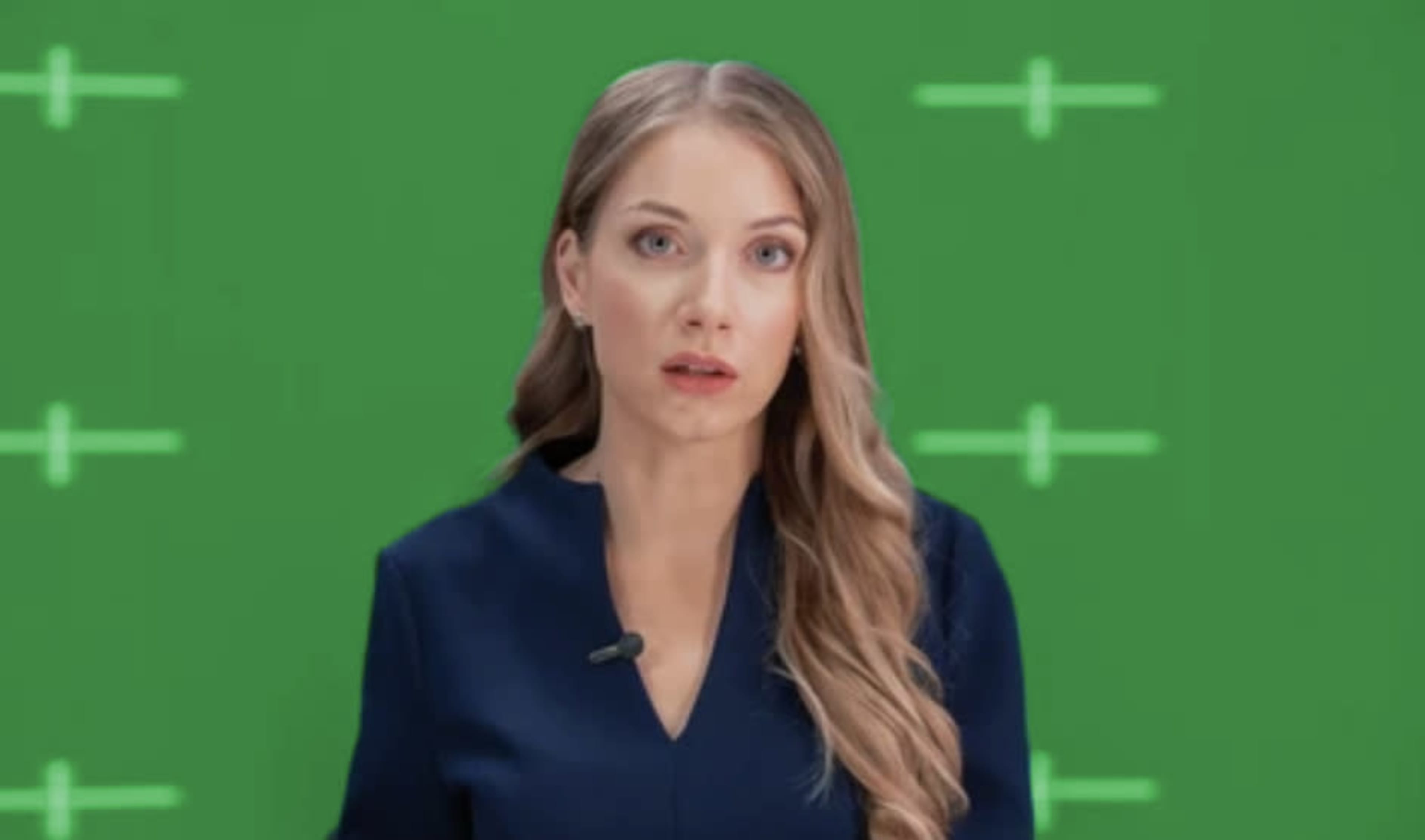 A woman with long, wavy hair stands in front of a green screen, wearing a dark outfit and looking directly at the camera.
