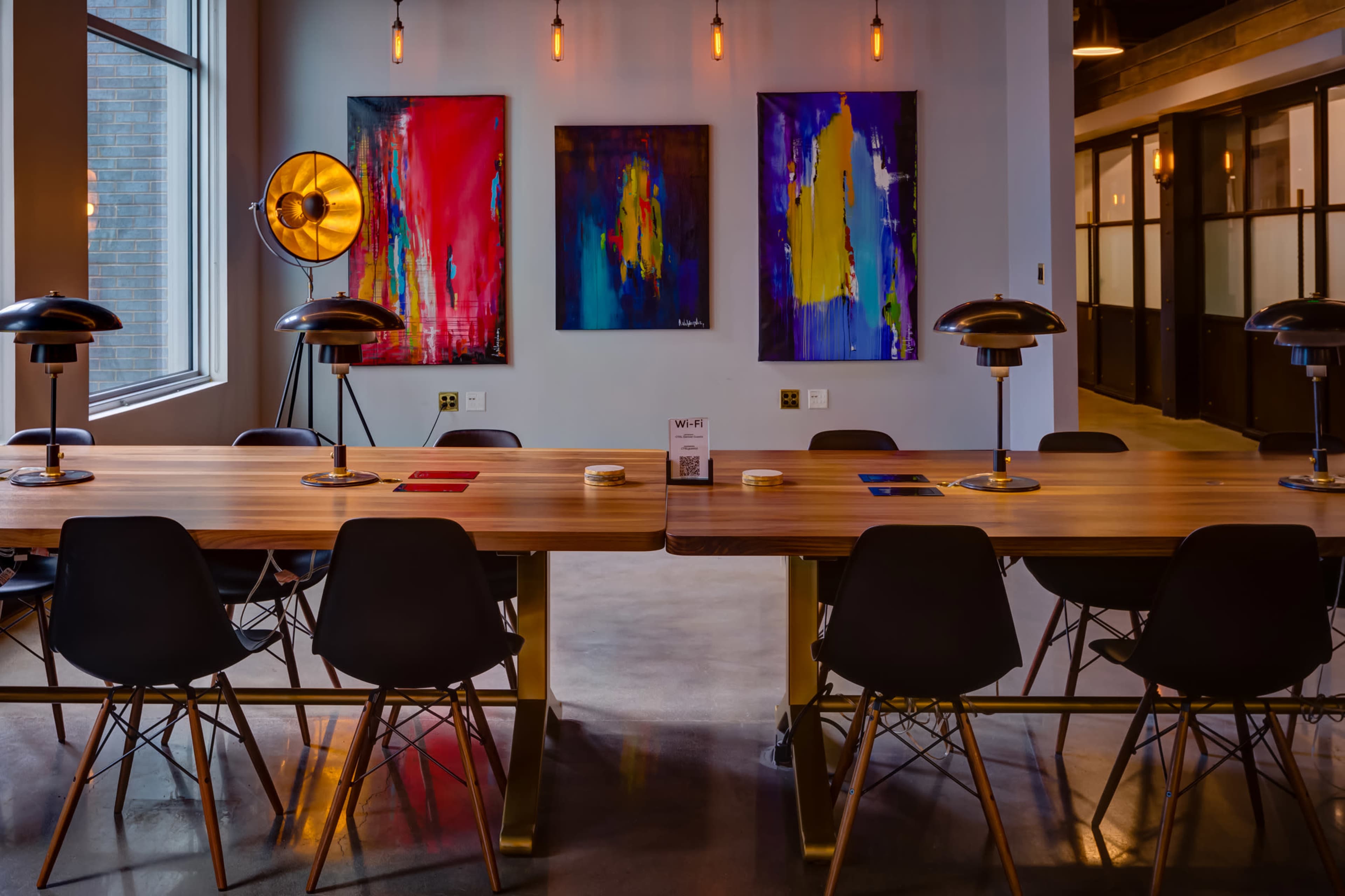 A long wooden table with black chairs is positioned in a modern room featuring colorful abstract paintings on the walls and stylish light fixtures overhead.