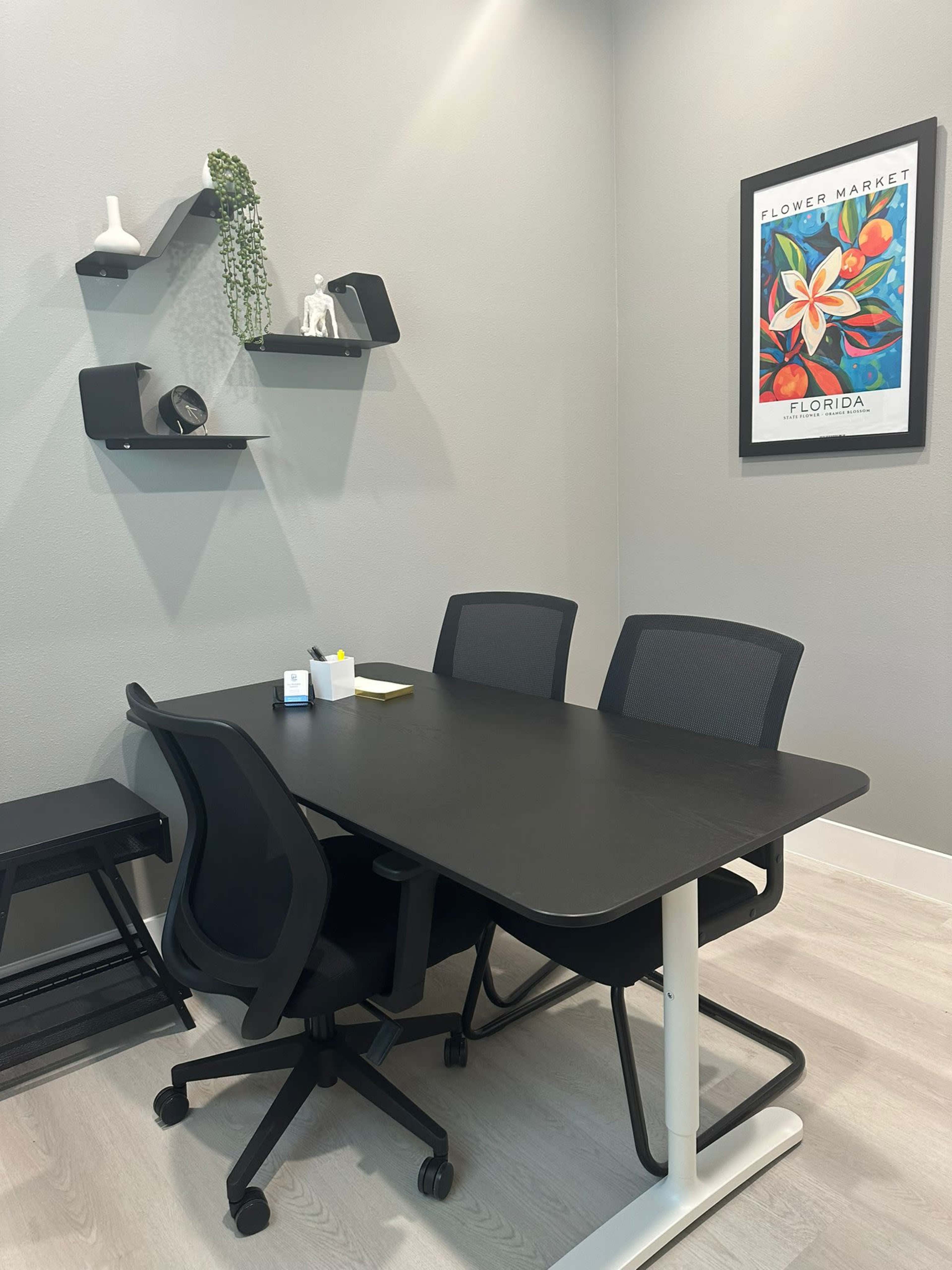 A modern conference room features a black table with three ergonomic chairs, a wall shelf displaying decorative items, and a framed artwork.