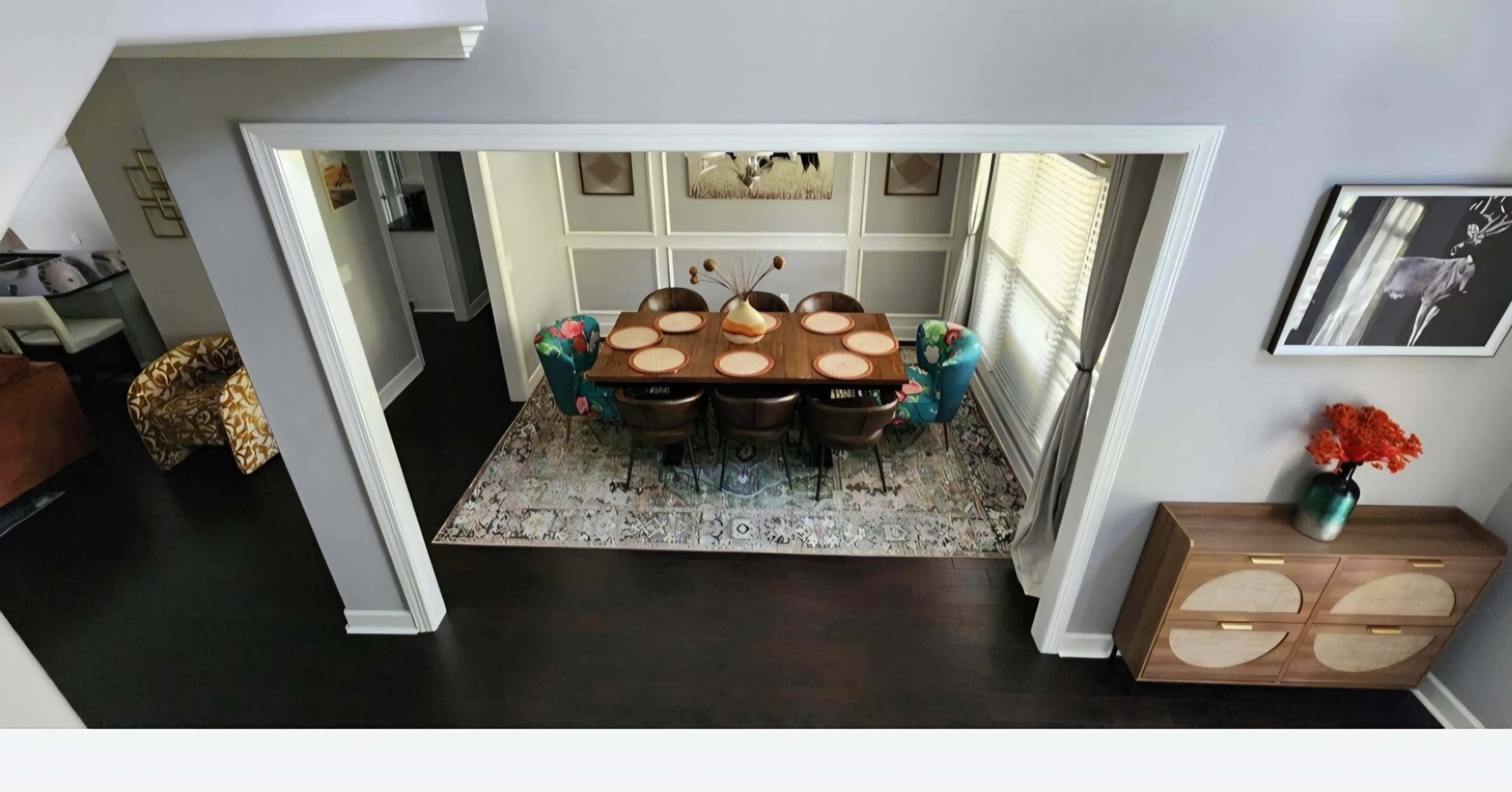 The image shows a dining room featuring a long table set for a meal, surrounded by eight chairs, with decorative accents and a patterned rug underneath.
