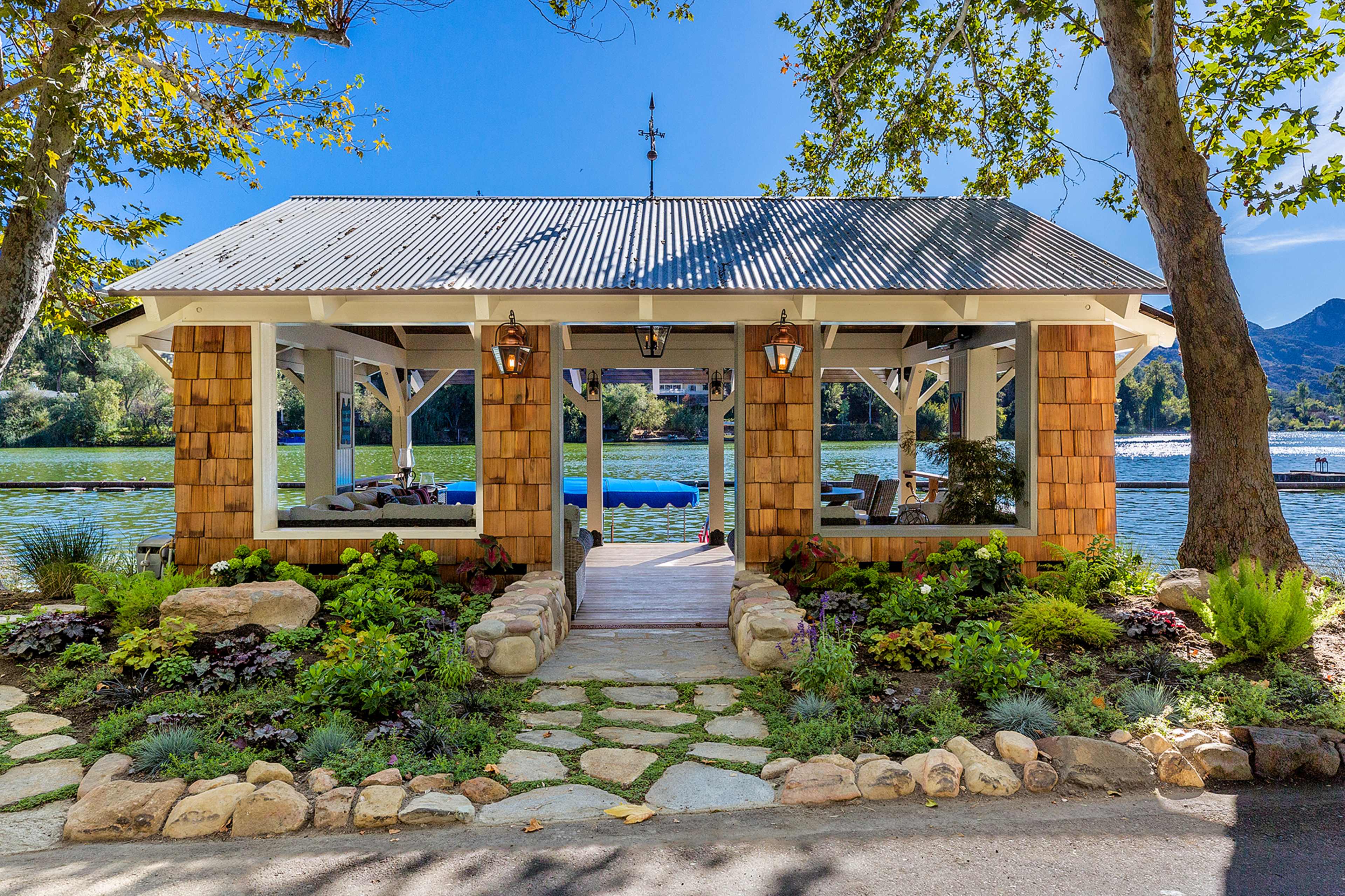 A lakeside gazebo with a stone walkway, surrounded by landscaped gardens and trees.