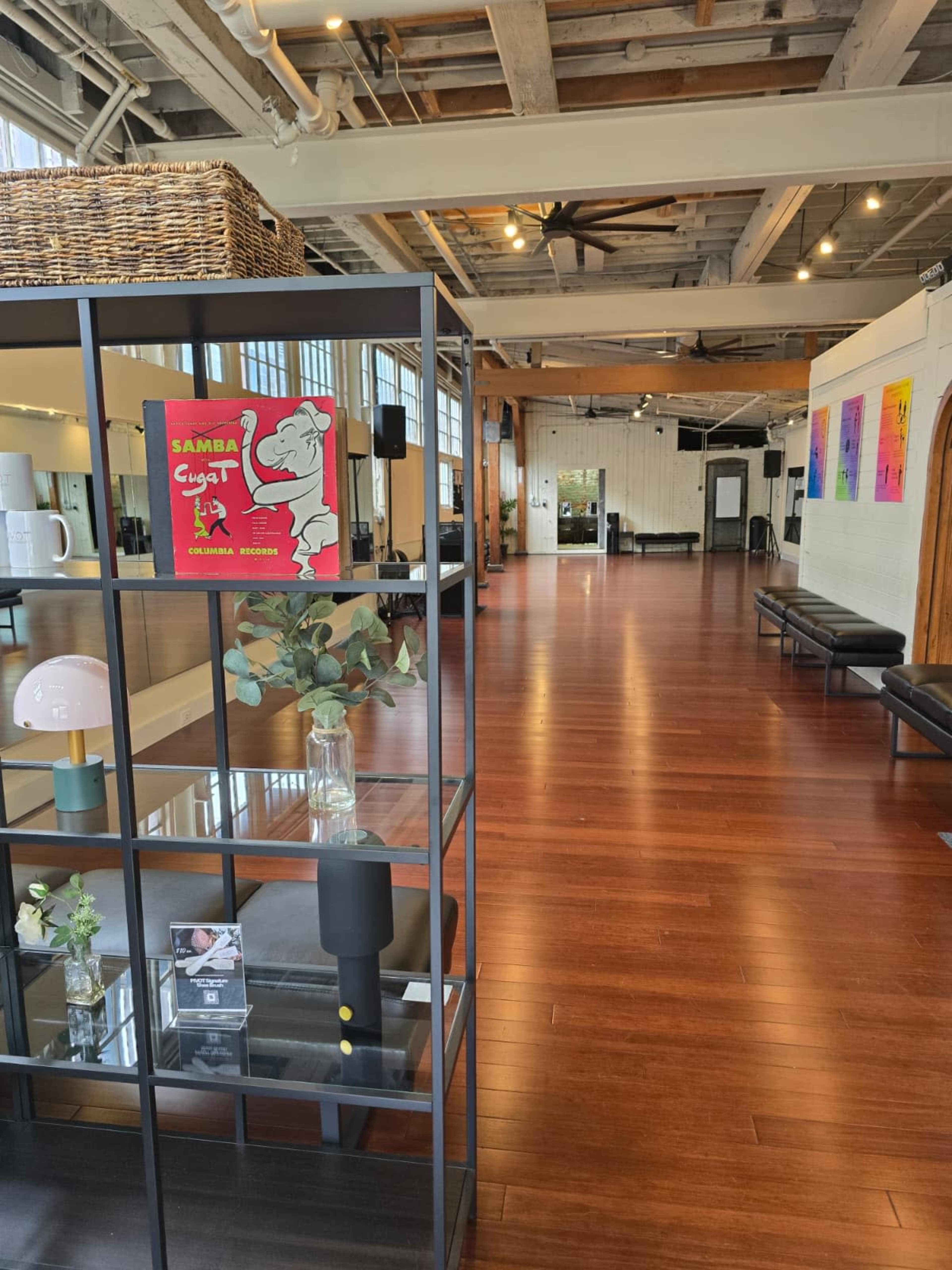 The image shows an expansive, well-lit interior space featuring polished wooden floors, a display shelf with decorative items, and various artworks on the walls.