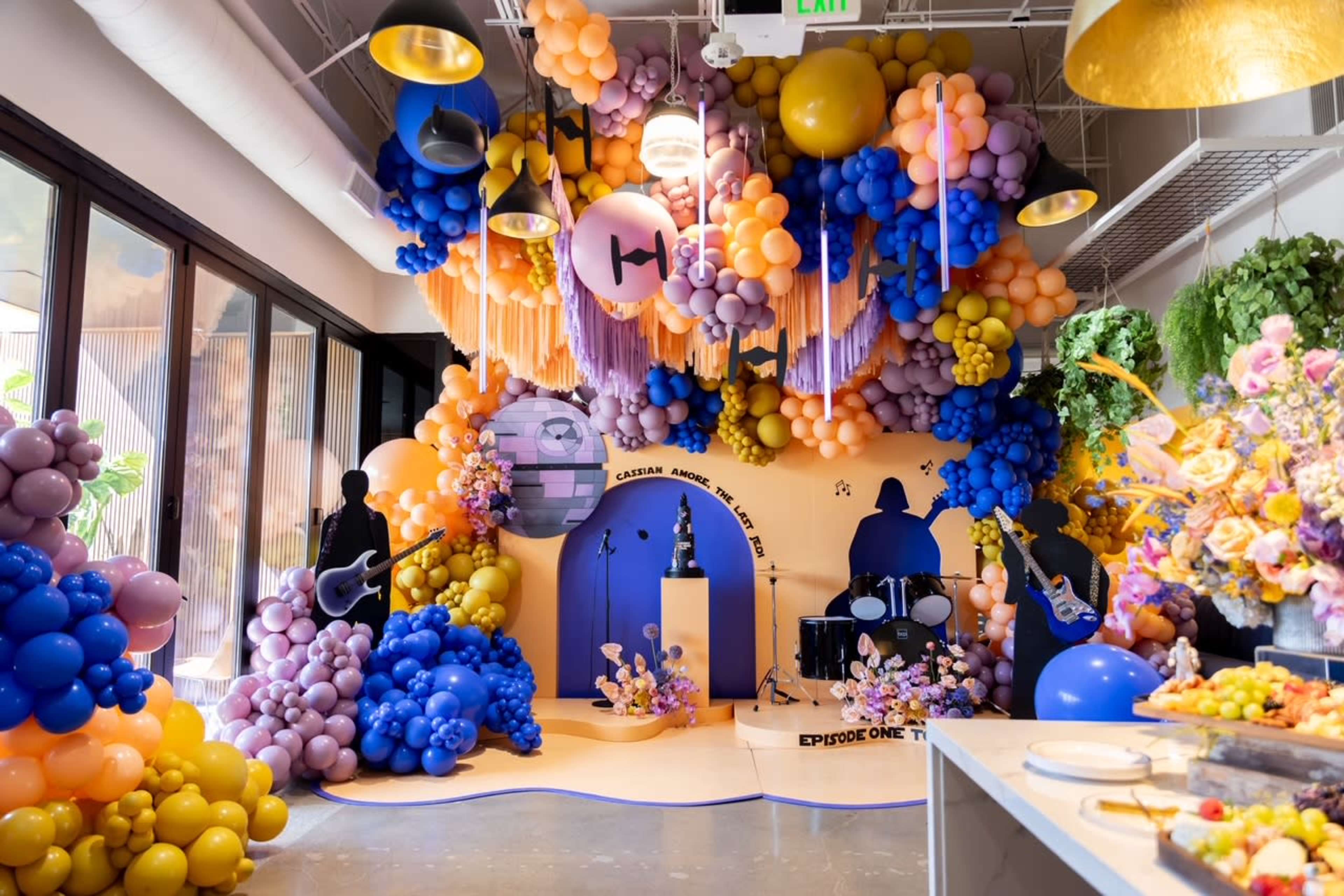 The image shows a colorful balloon installation featuring various shades of purple, yellow, and orange, arranged around a stage with silhouettes of musicians and a drum set in a modern space.