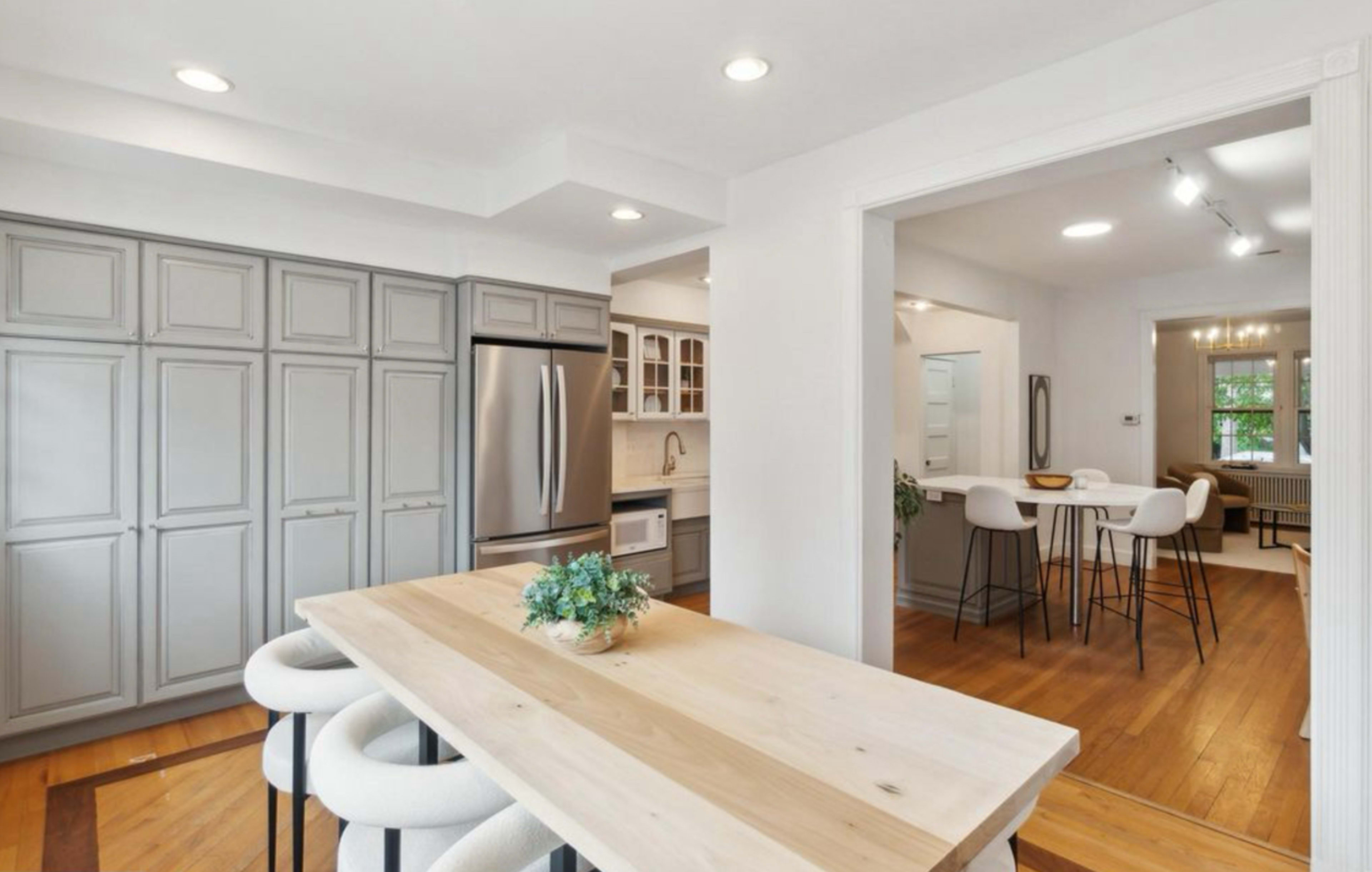 The image shows a modern kitchen with gray cabinets, a central wood dining table, and an open layout connecting to a living area.