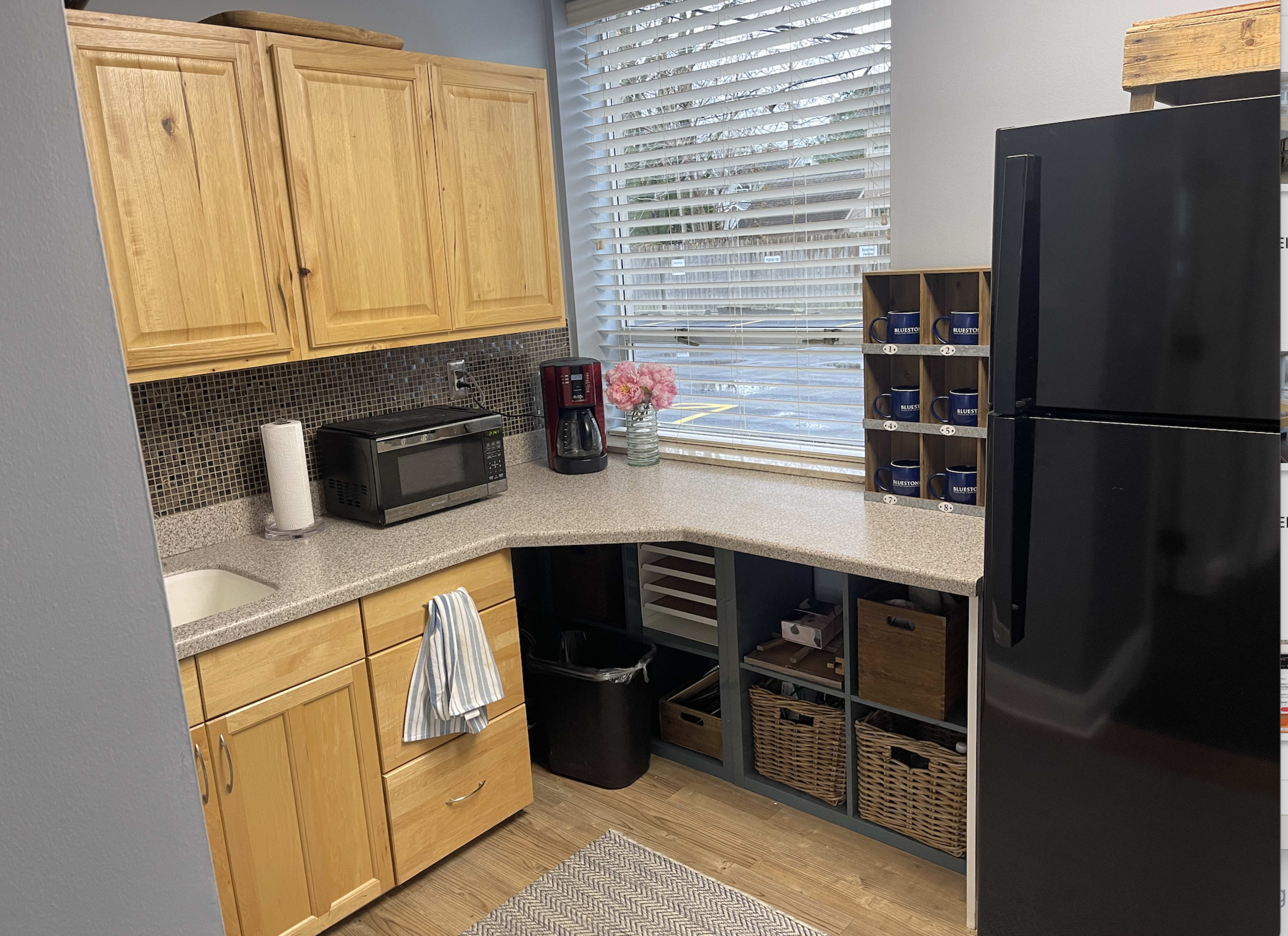 A compact kitchen features wooden cabinets, a countertop with a sink, a microwave, a coffee maker, and a black refrigerator, alongside a window with blinds and decorative shelving.