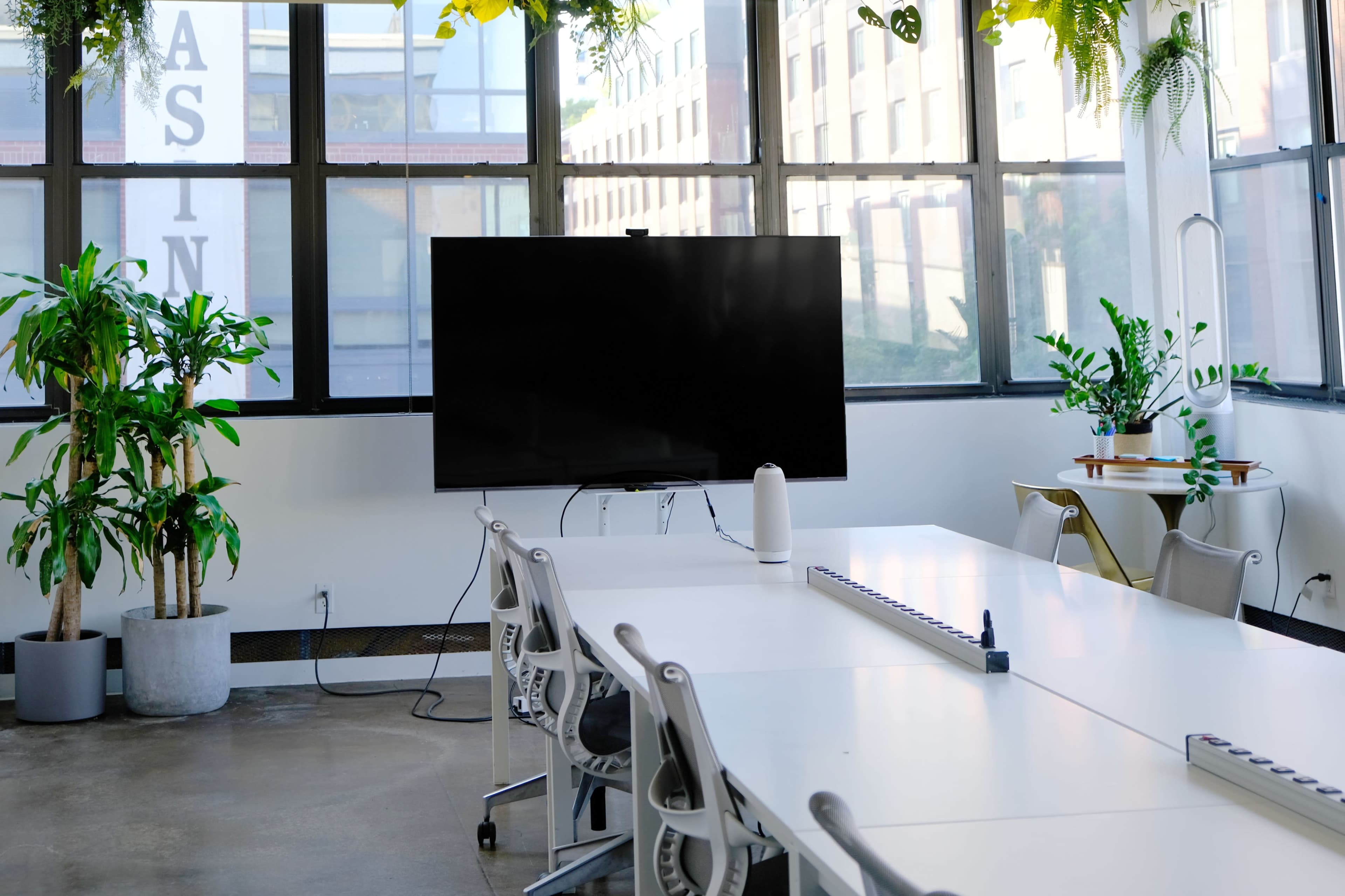 A modern conference room features a large screen, white tables, and several potted plants.