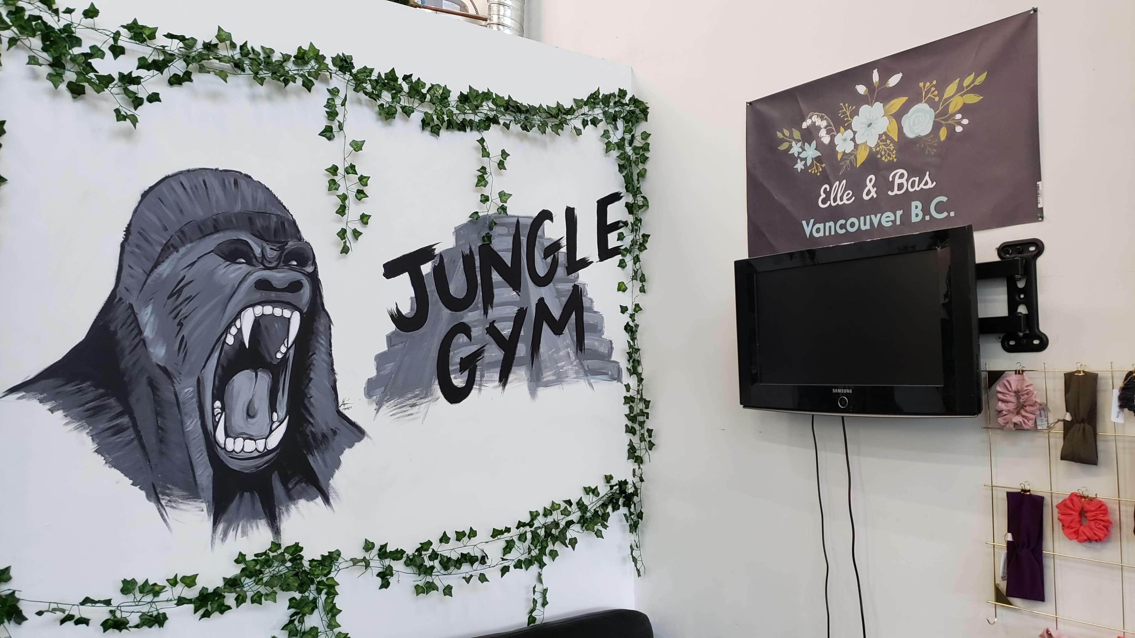 A mural of a roaring gorilla with the words "Jungle Gym" next to it, adorned with green vines, alongside a wall-mounted television and a decorative sign for "Elle & Bas" in Vancouver, B.C.