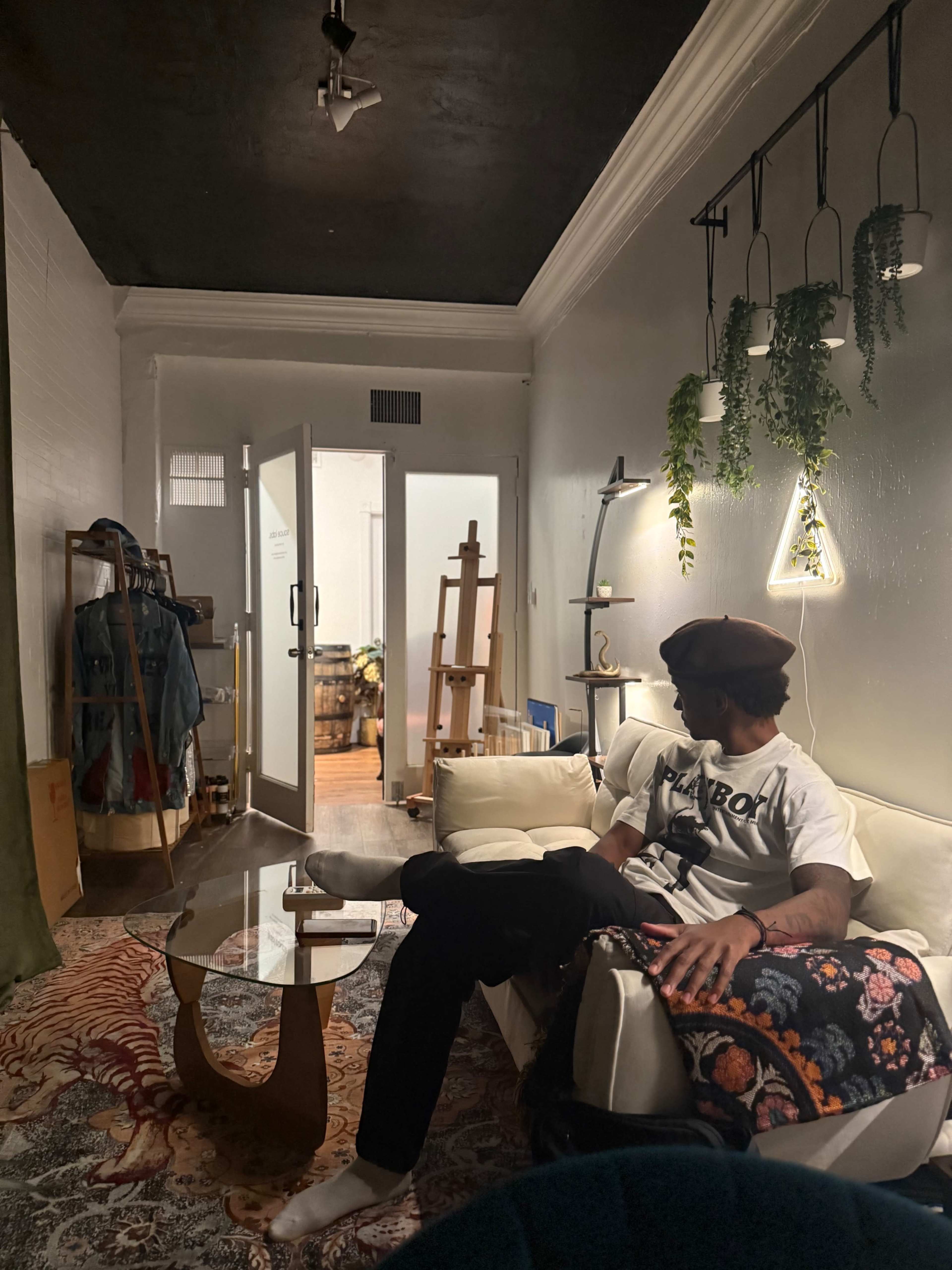 A person sits on a couch in a decorated living room with plants hanging from the ceiling and an easel visible in an adjacent room.