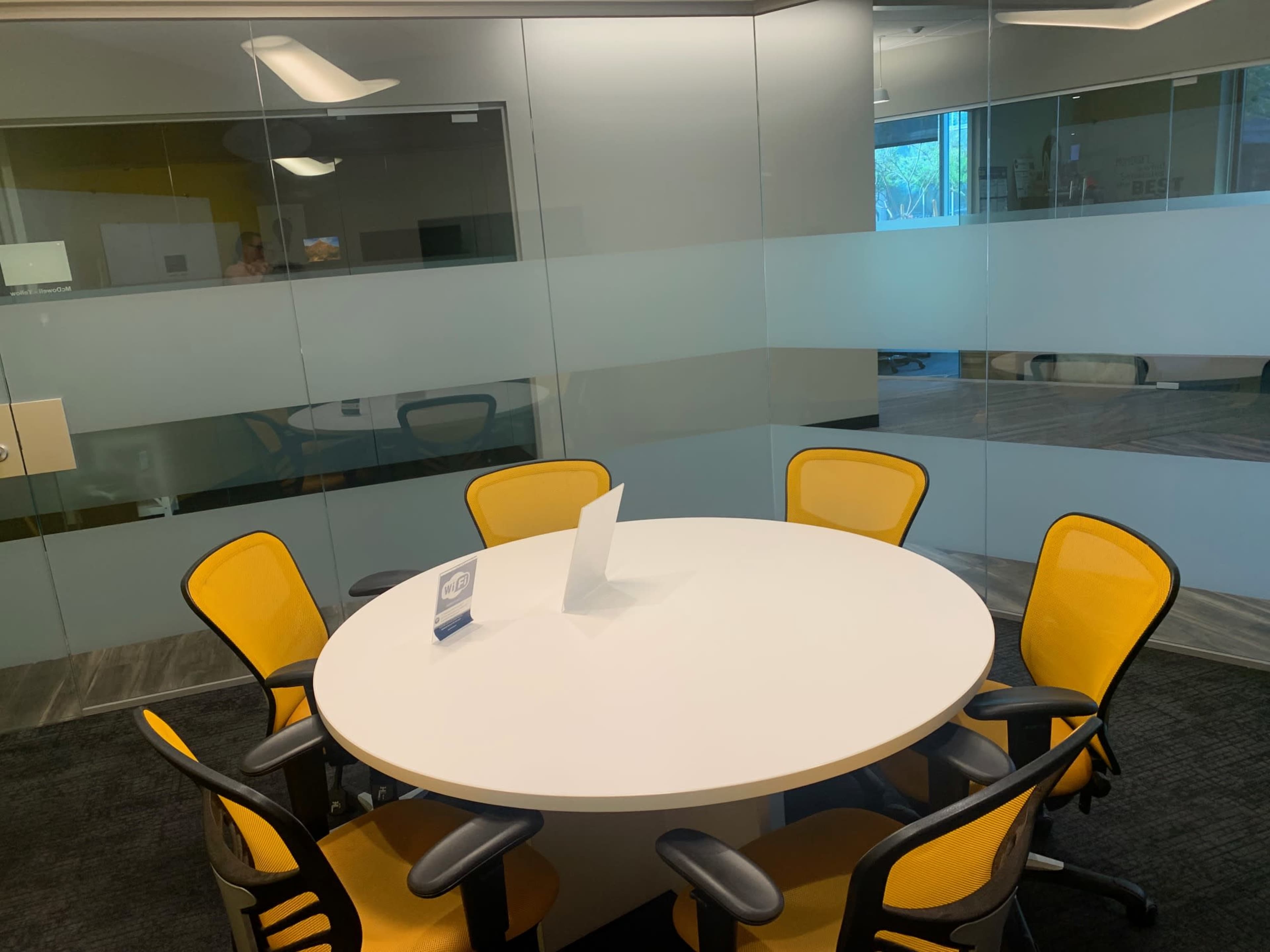 A round table with a white surface is surrounded by six yellow chairs in a glass-walled meeting room.