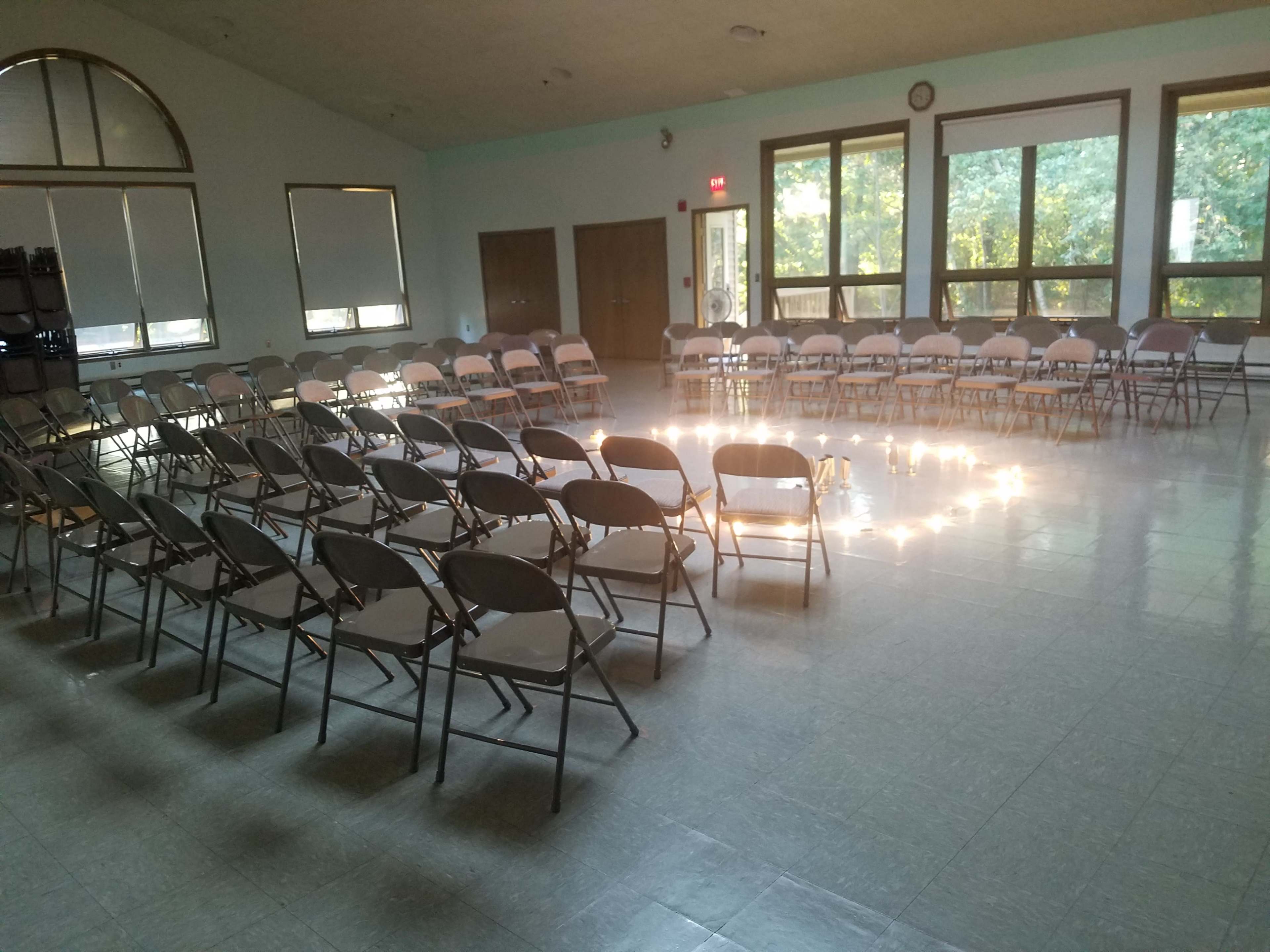 A room is arranged with rows of folding chairs facing a circular formation in the center, illuminated by natural light from large windows.