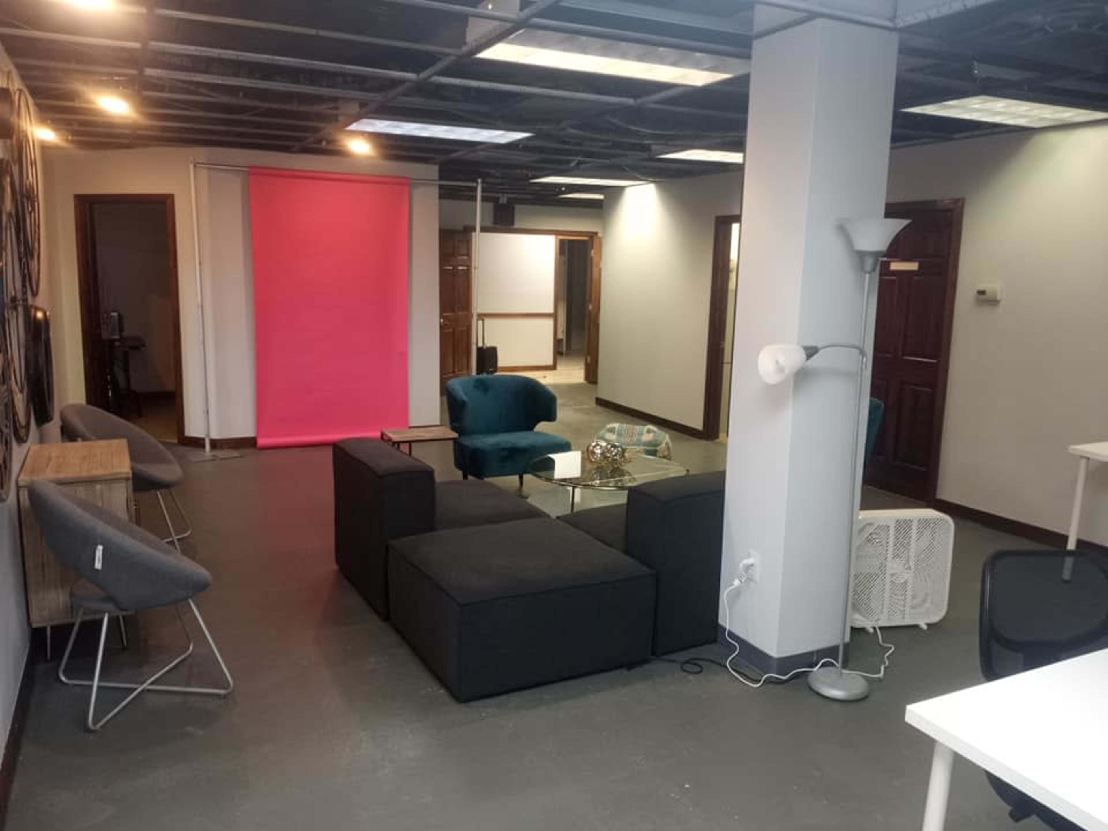 The image shows a modern office space featuring a seating area with a dark sofa, light blue chairs, a glass coffee table, and a bright pink backdrop.