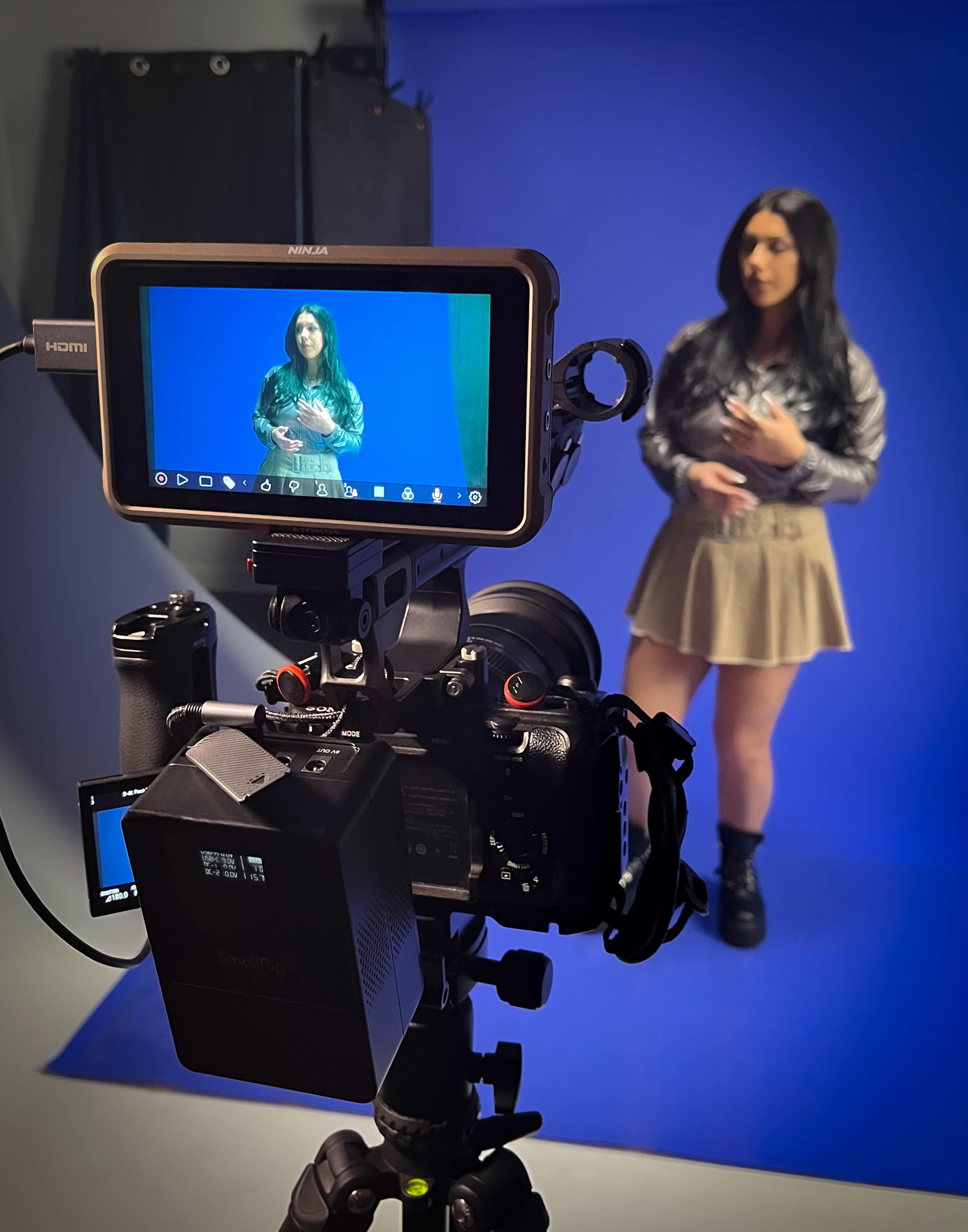 A camera is set up on a tripod in front of a blue backdrop, displaying a woman holding an object in a studio.