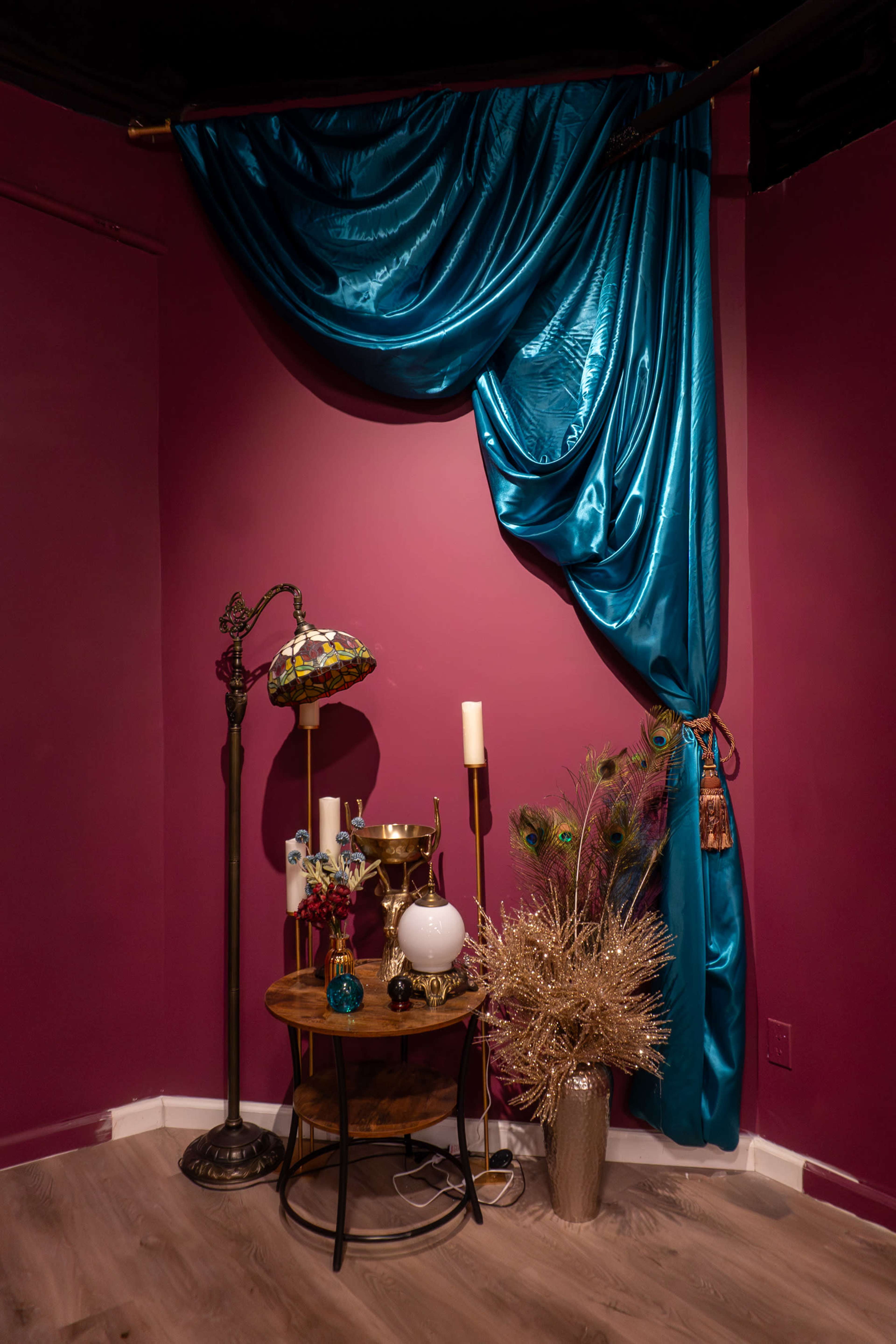 A corner of a room features a vibrant purple wall, a flowing turquoise fabric drapery, a vintage lamp, decorative candles, and a floral arrangement in a vase.