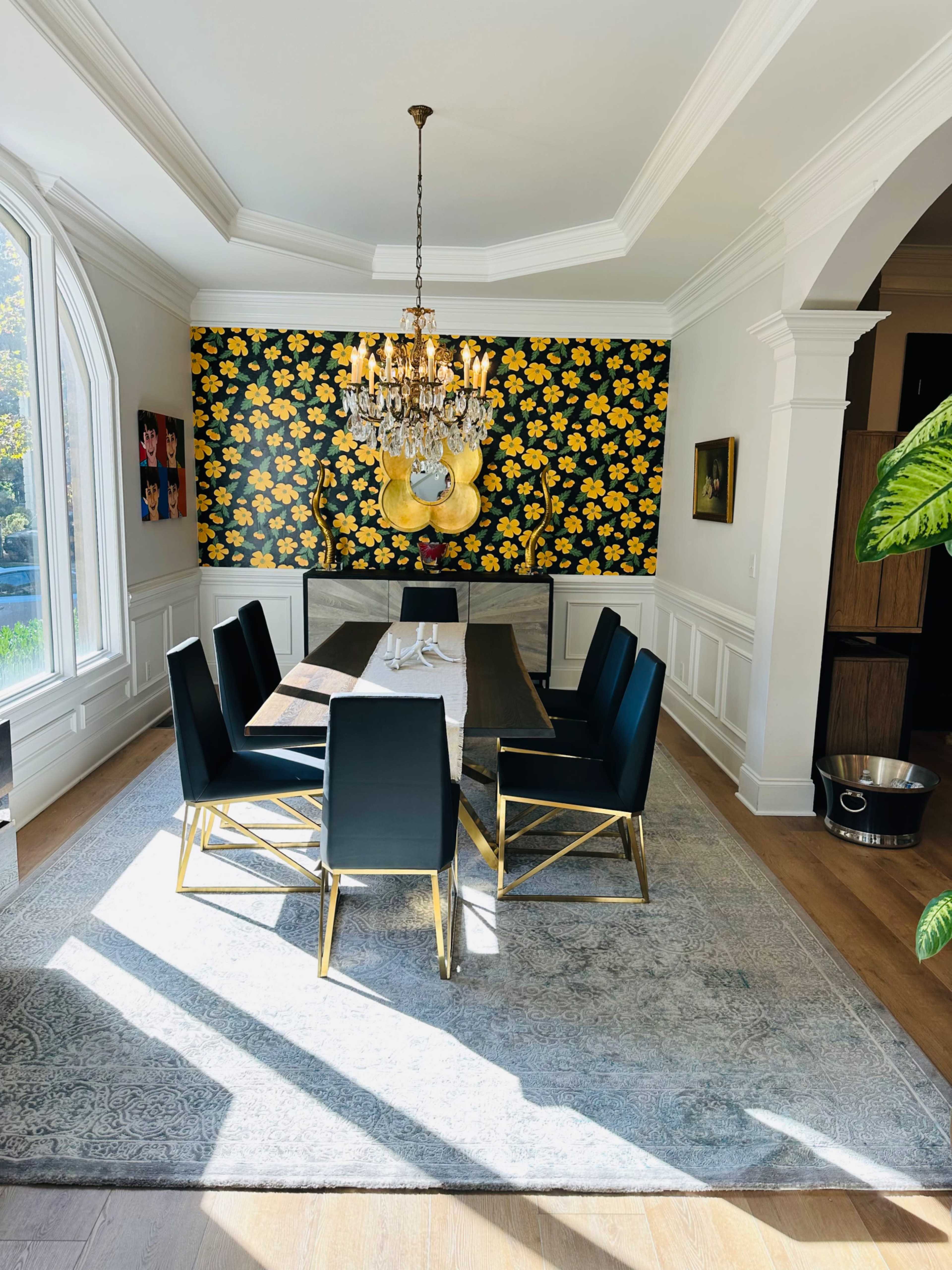 The image shows a modern dining room featuring a black table surrounded by six black chairs, a chandelier overhead, and a floral-patterned accent wall.