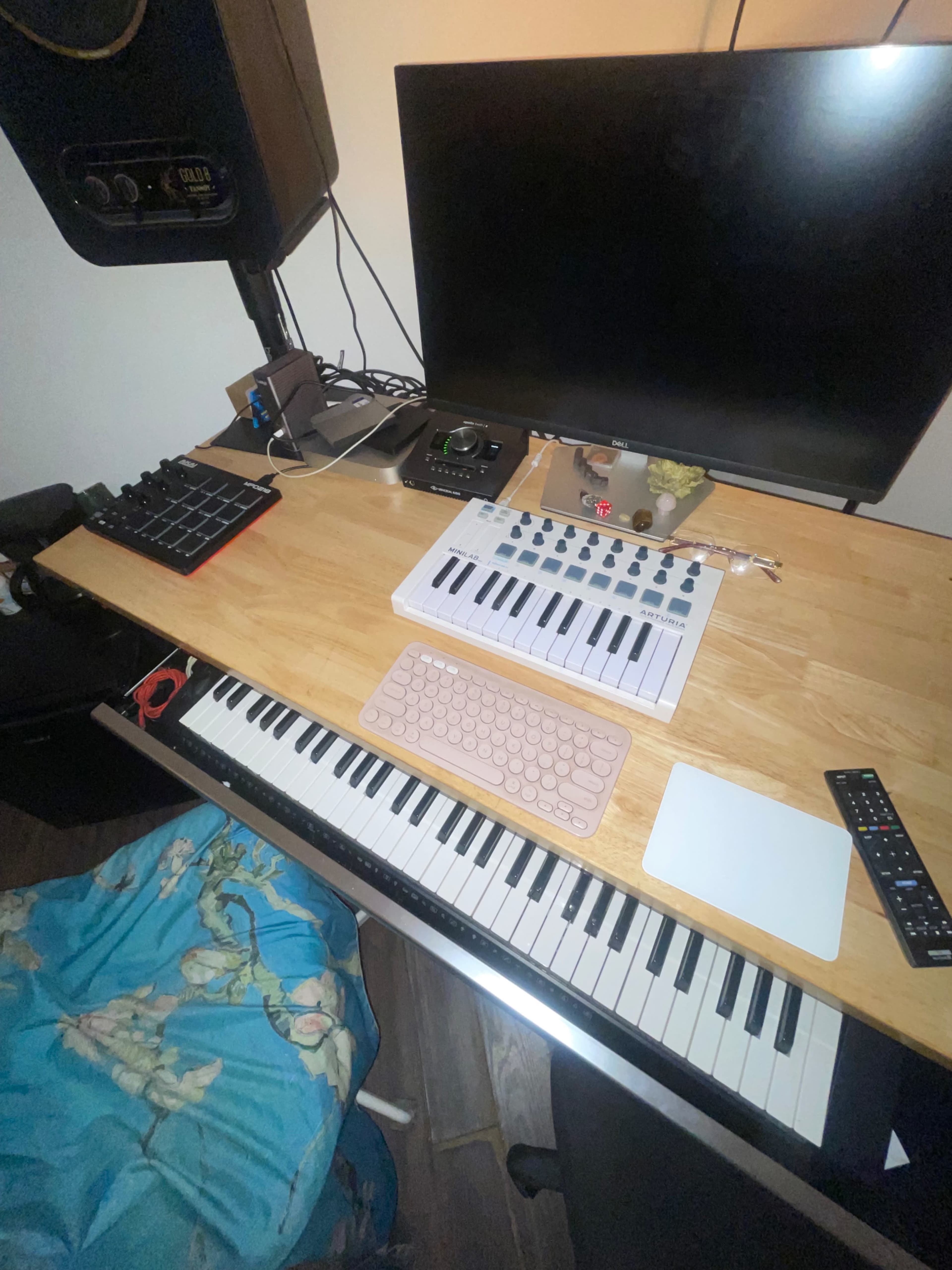 A music production workstation features a keyboard, a laptop, a monitor, and various equipment on a wooden desk.