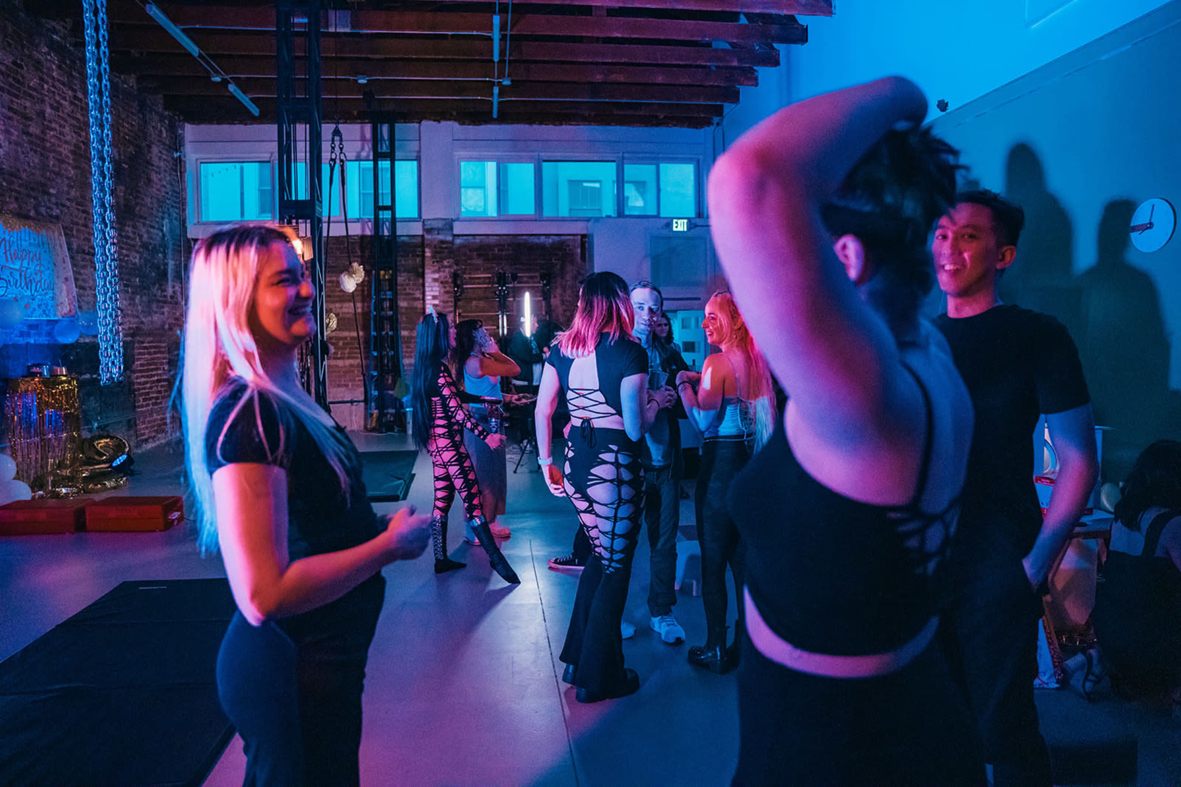 A group of people in black clothing socialize in a brightly lit studio with exposed brick walls and vibrant lighting.