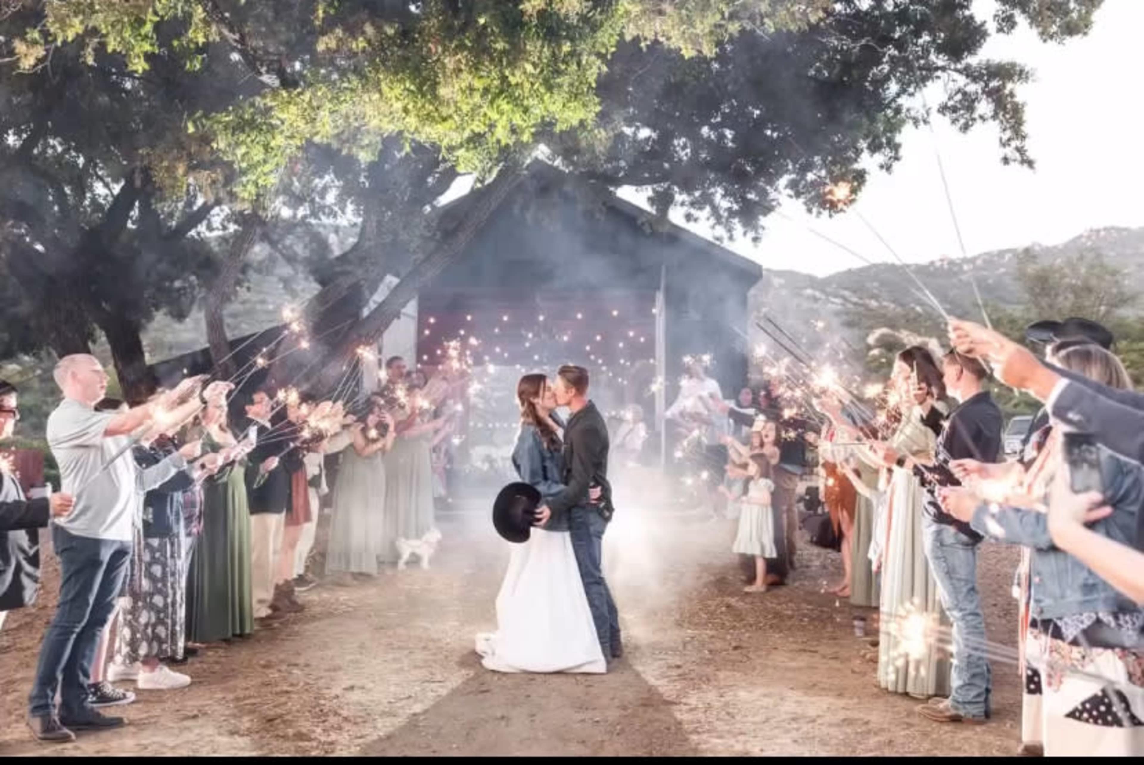 A couple kisses in front of a rustic building as guests hold sparklers and celebrate their wedding.