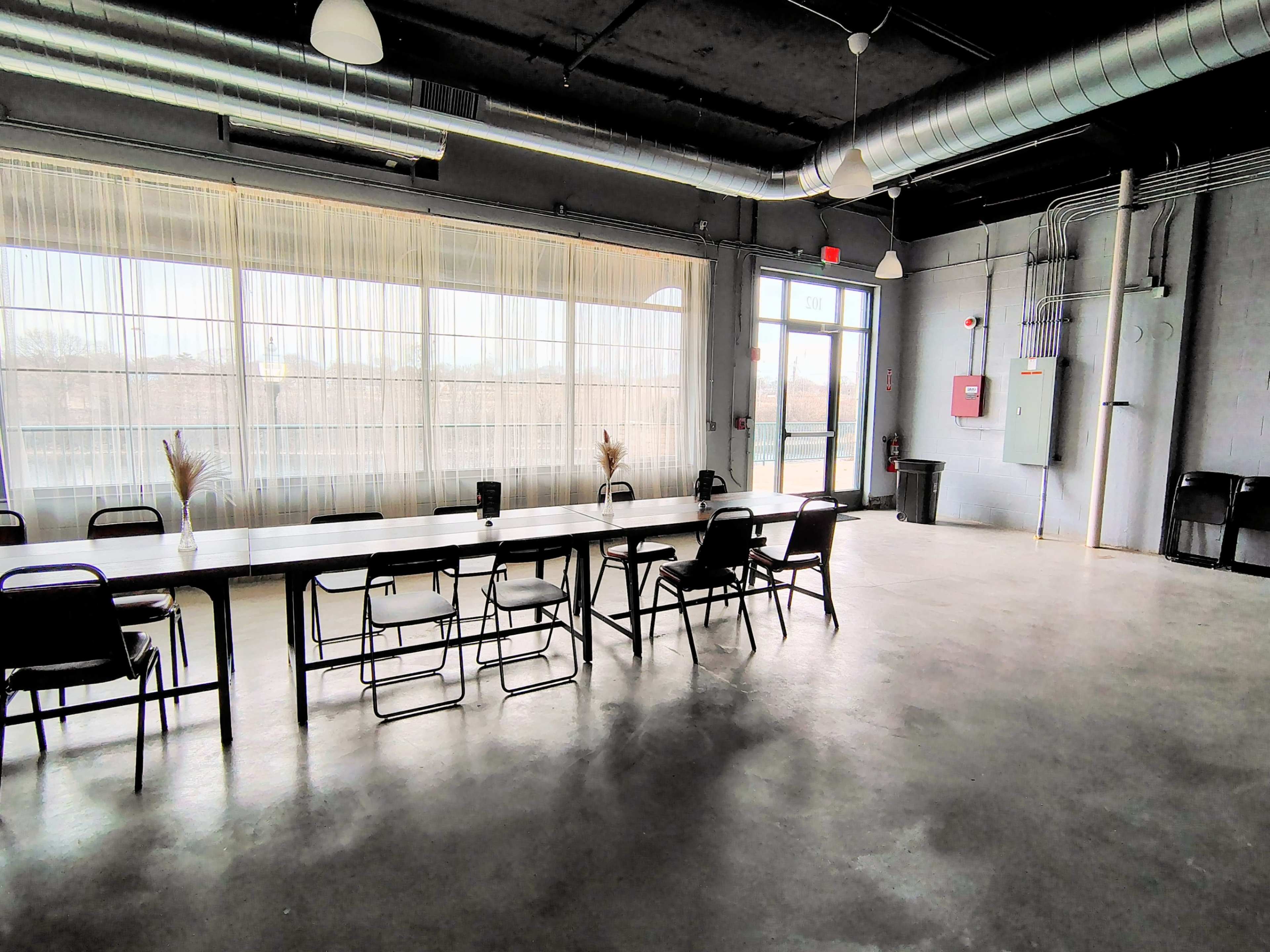 The image shows a spacious, modern room with a long table and chairs, large windows draped with sheer curtains, and industrial-style elements such as exposed pipes.