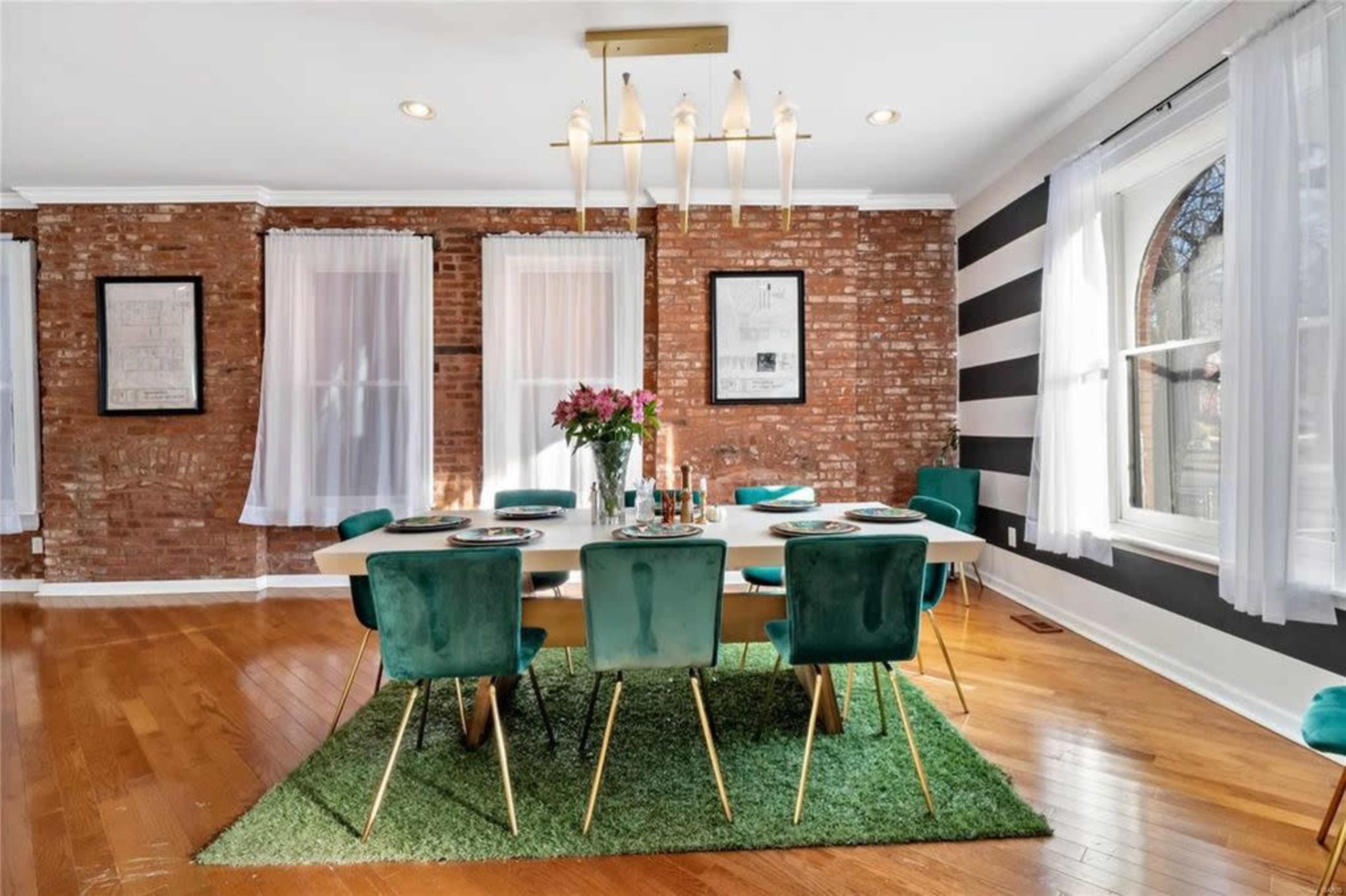 A dining room features a white table surrounded by green chairs on a rug, with exposed brick walls and large windows.