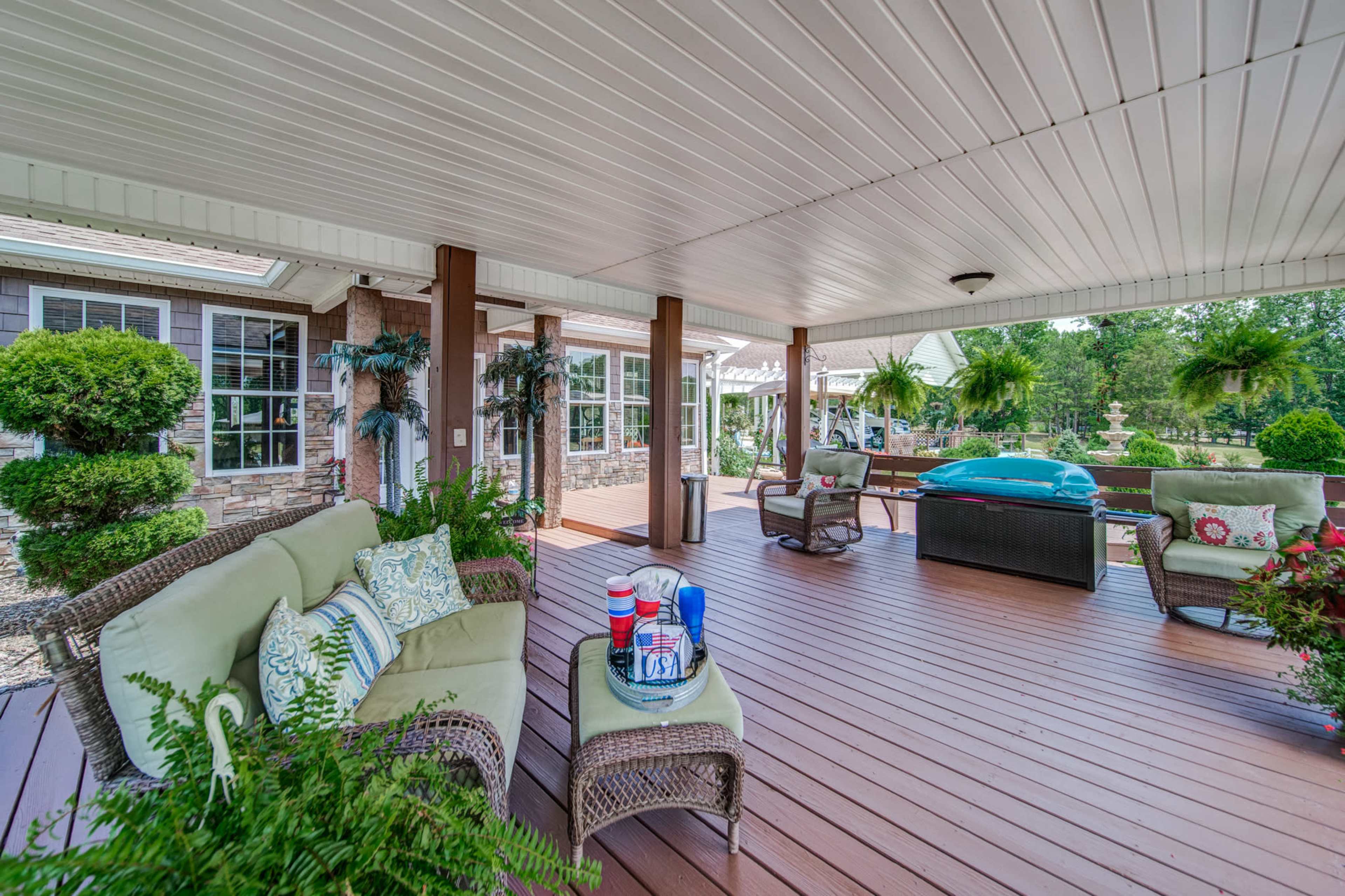 The image shows an outdoor covered patio with comfortable seating, potted plants, and a hot tub in a landscaped yard.