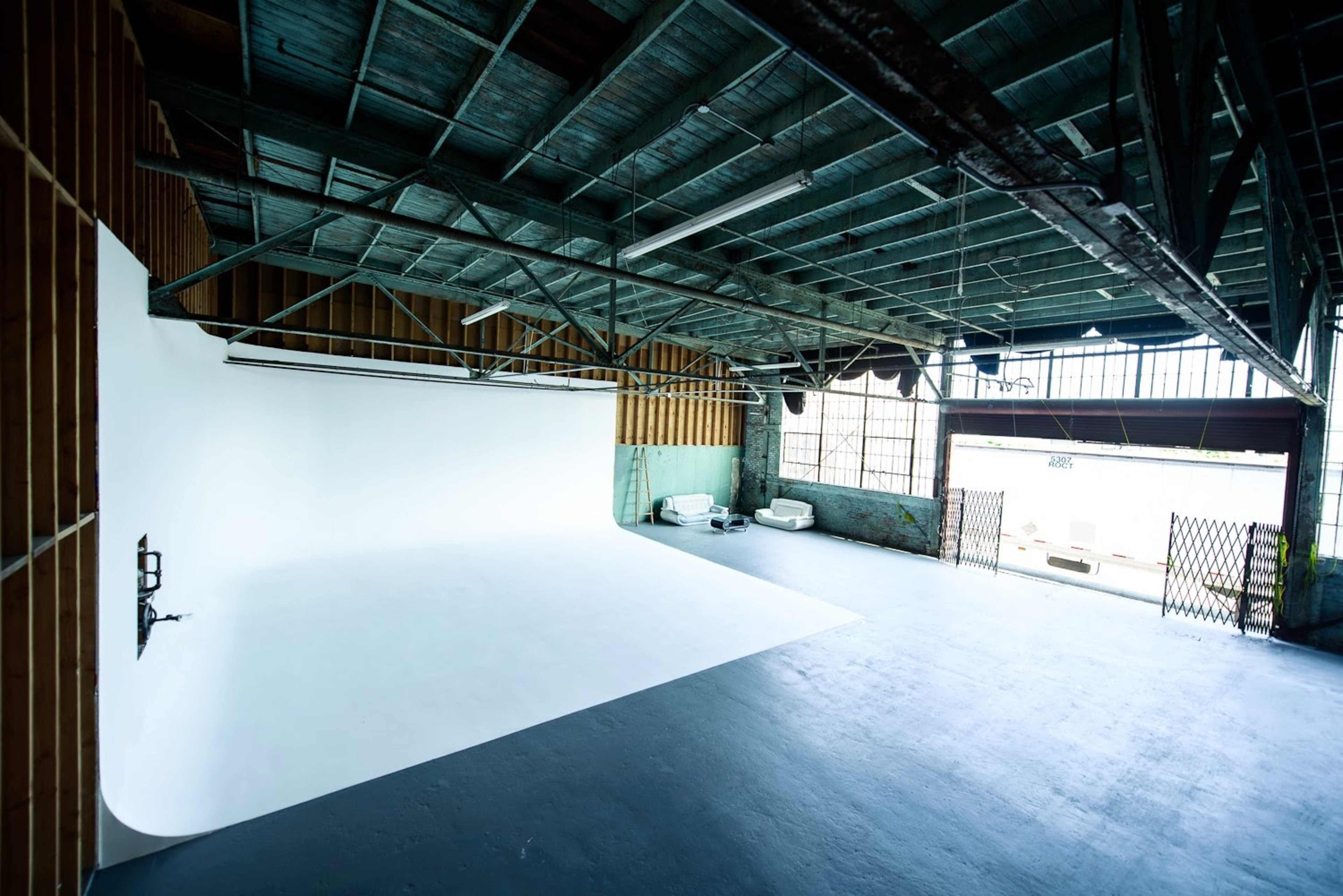 A spacious photography studio with a large white backdrop, exposed beams, and large windows letting in natural light.