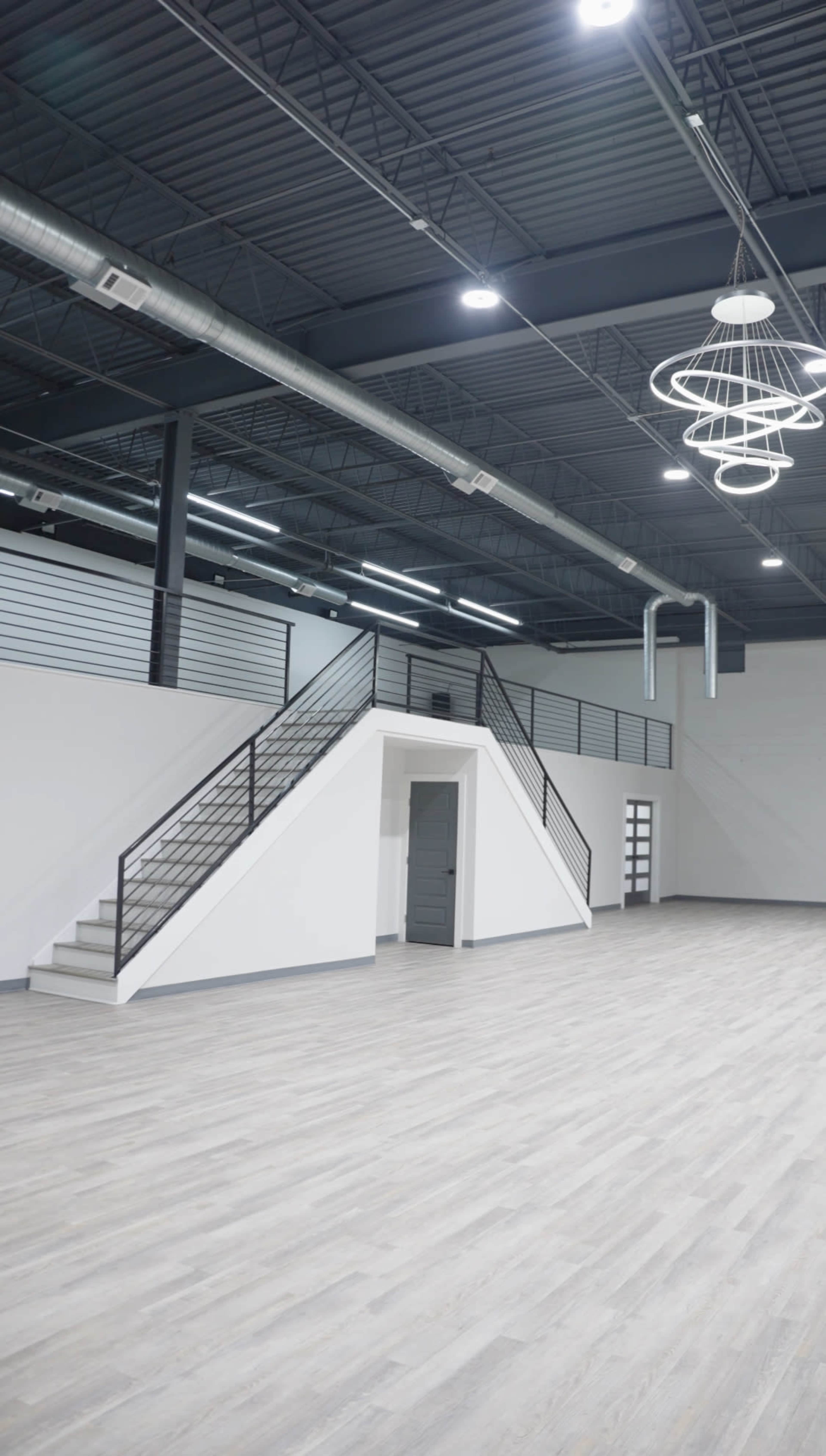 The image shows a spacious, modern interior of a building with a staircase leading to a second floor and large windows along the walls.