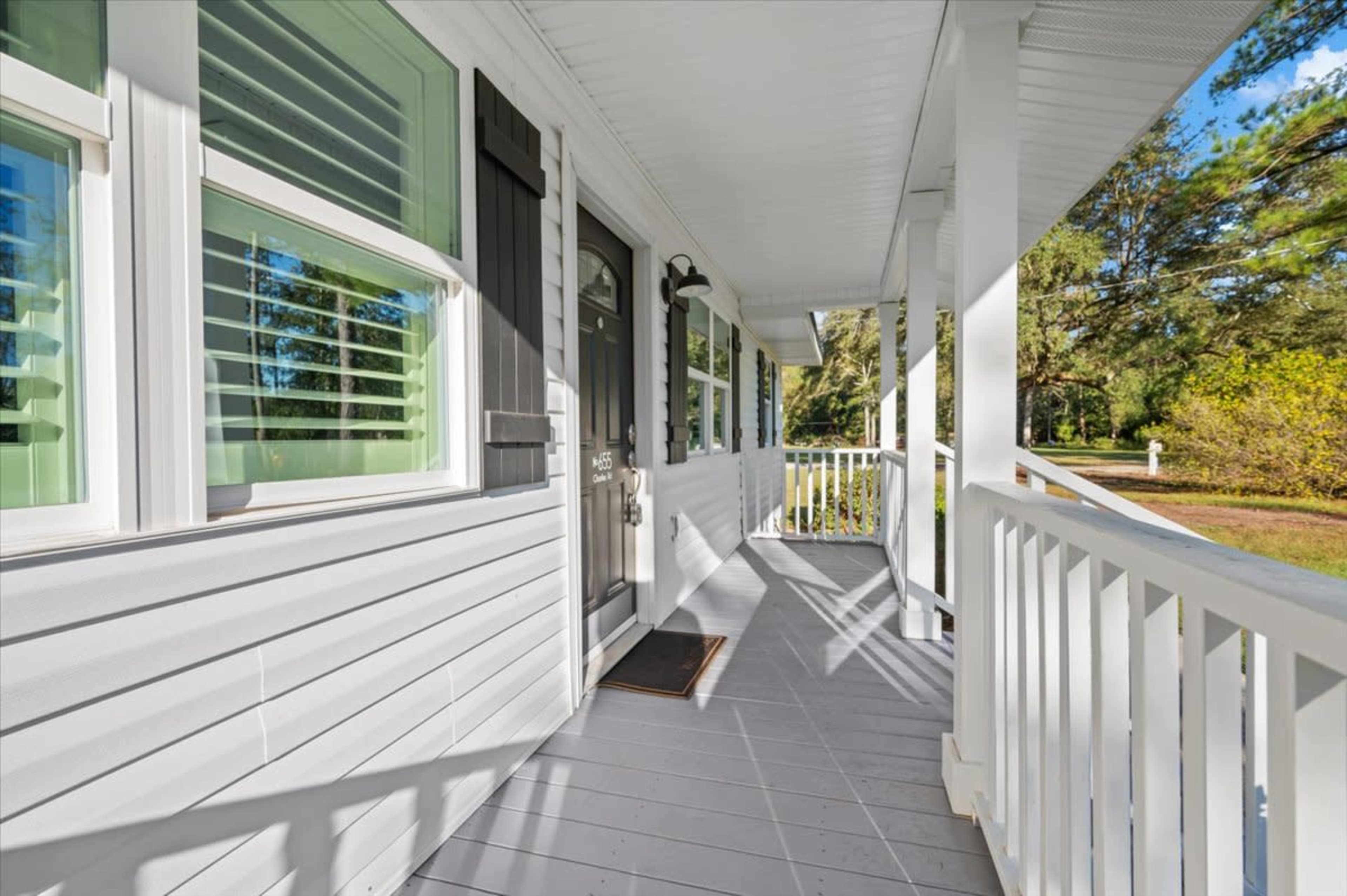 A porch area with a gray floor, white railings, and sunlight filtering through the trees, leading up to a front door framed by windows.
