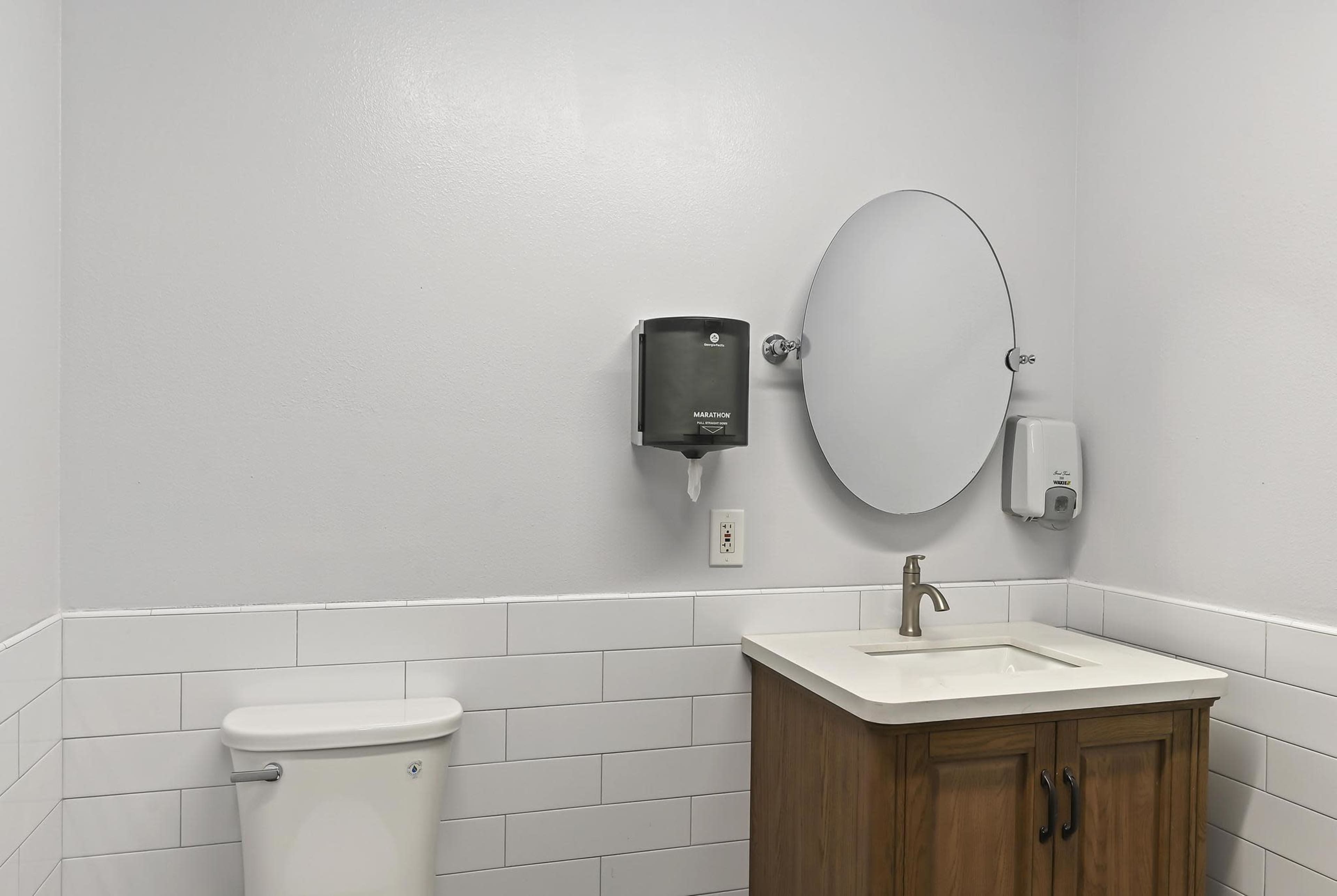 A clean bathroom featuring a toilet, a sink with a wooden cabinet, a mirror, and a soap dispenser on the wall.