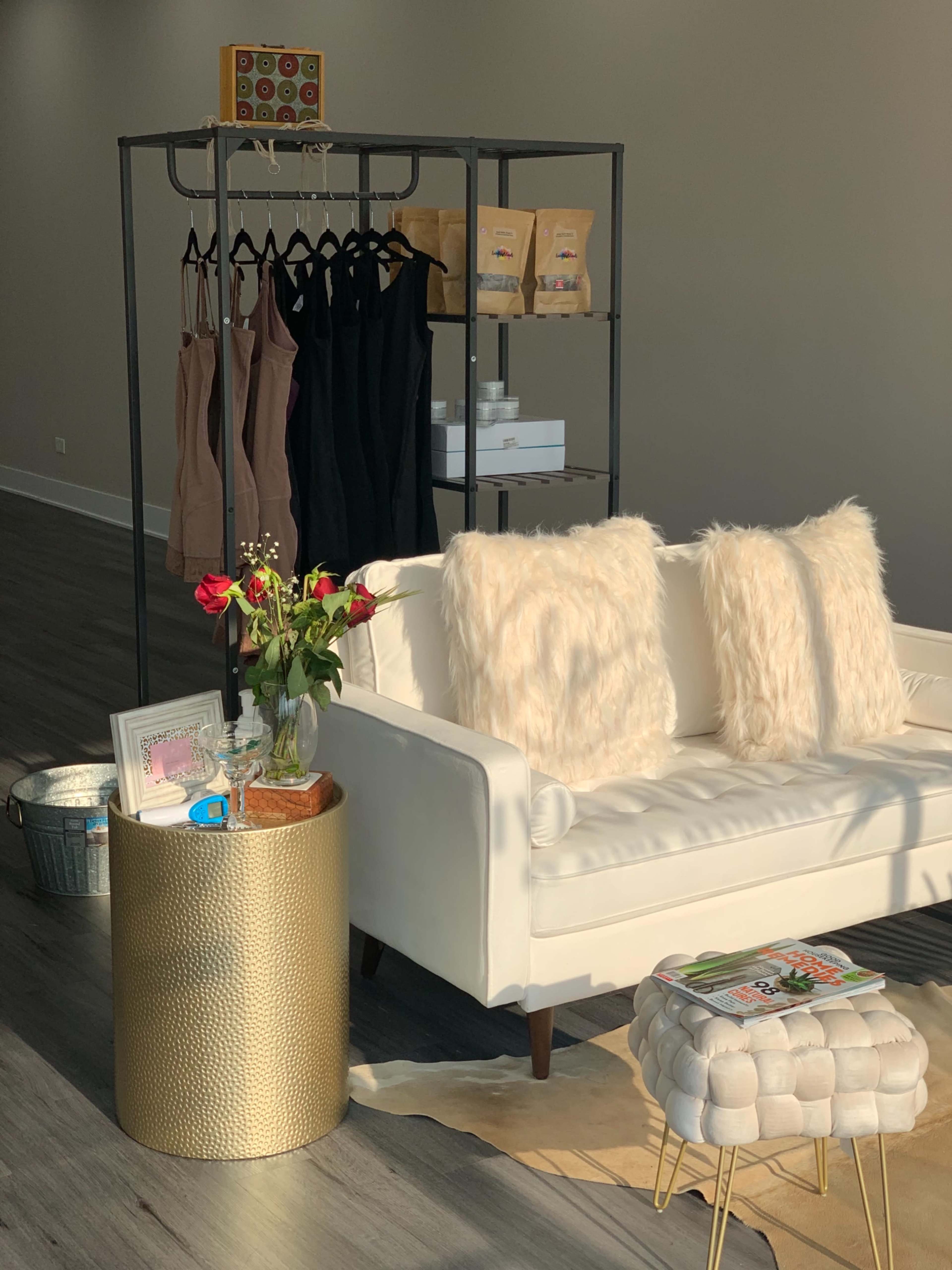 A white sofa with fluffy pillows sits next to a small gold table, while a rack holds garments and bags in the background.