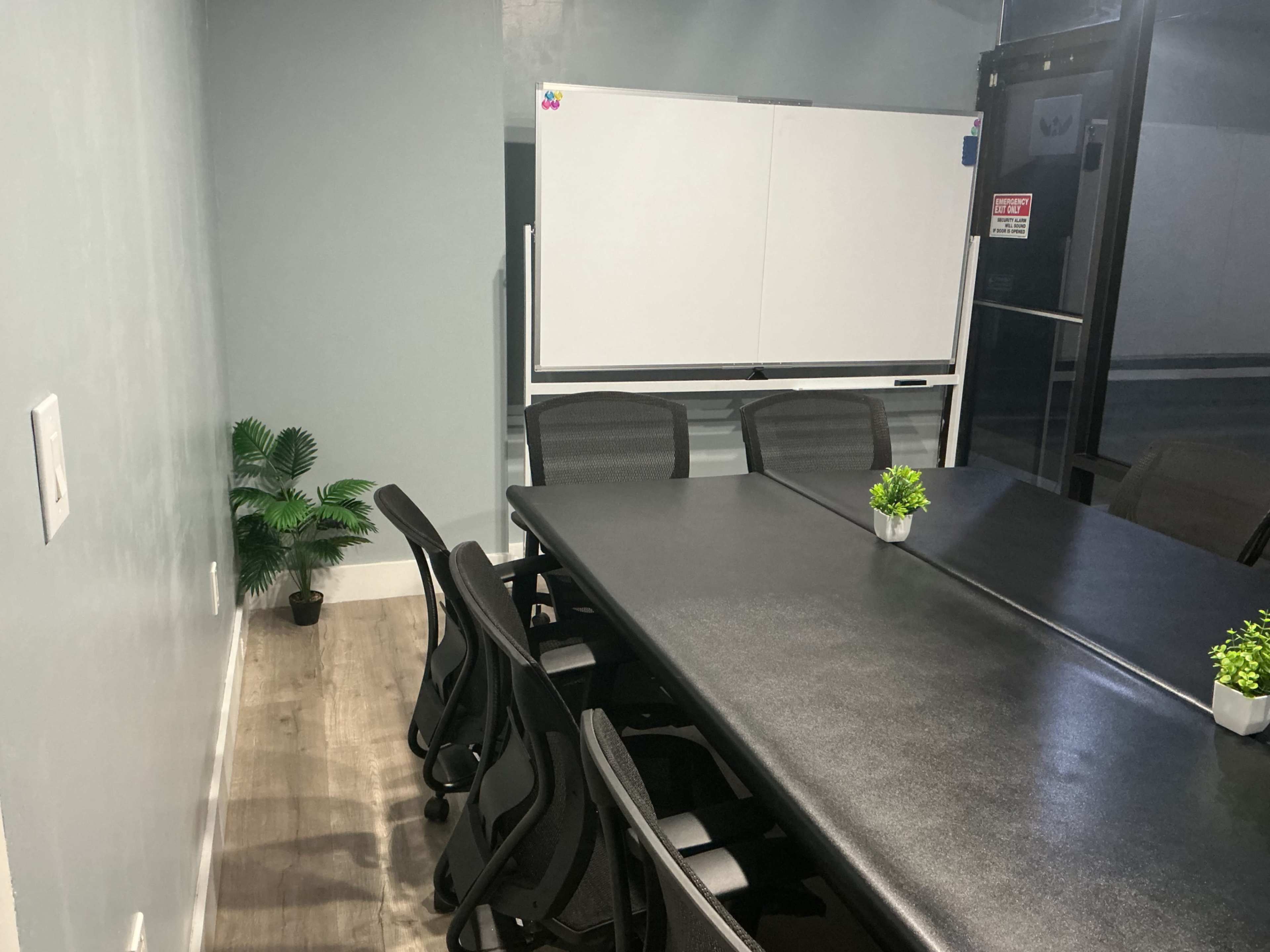A conference room features a large black table surrounded by ergonomic chairs, a whiteboard, and a small potted plant in the corner.