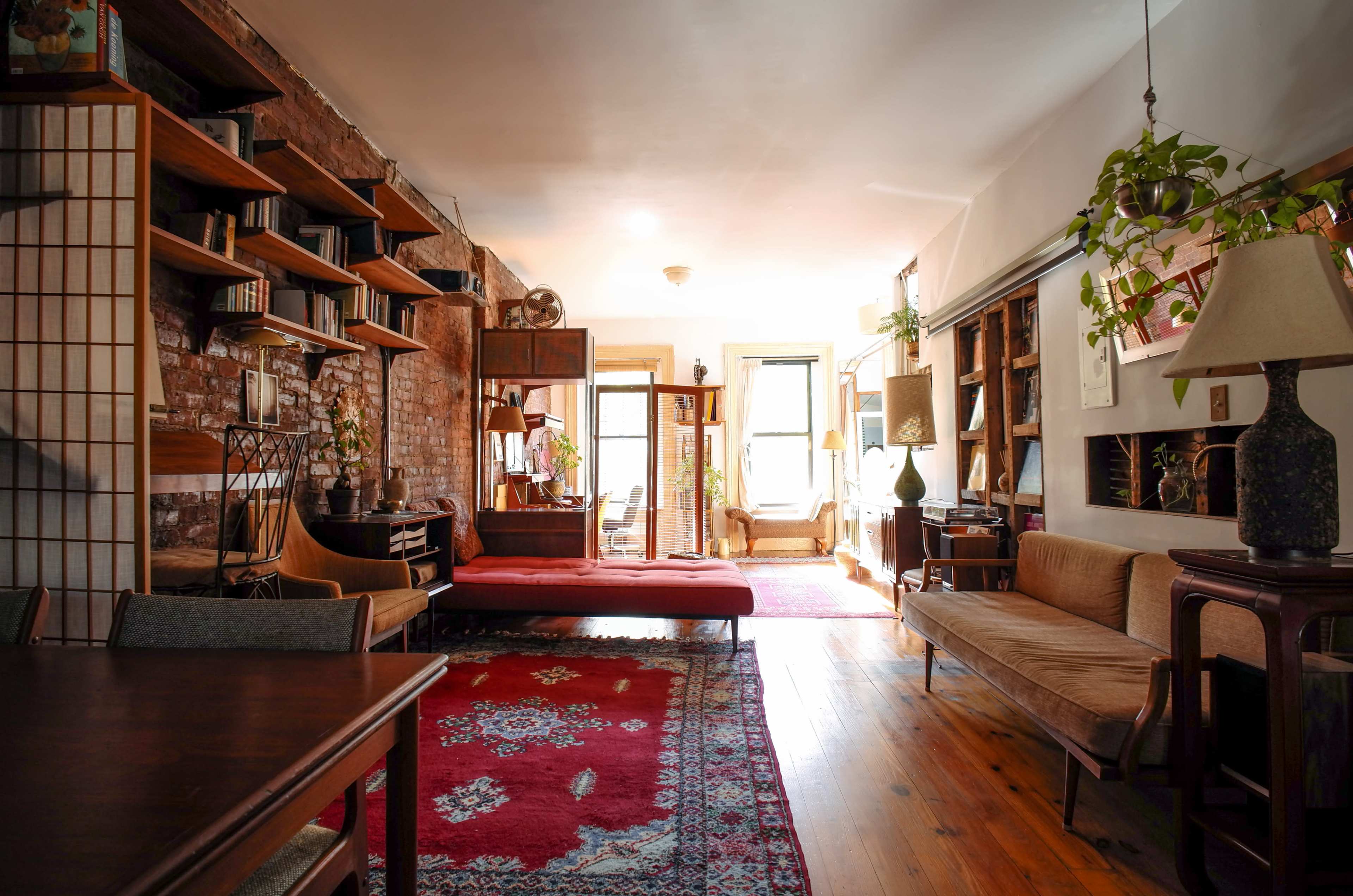 A cozy living room with a wooden floor, a patterned red rug, various seating options, and shelves filled with books and plants.