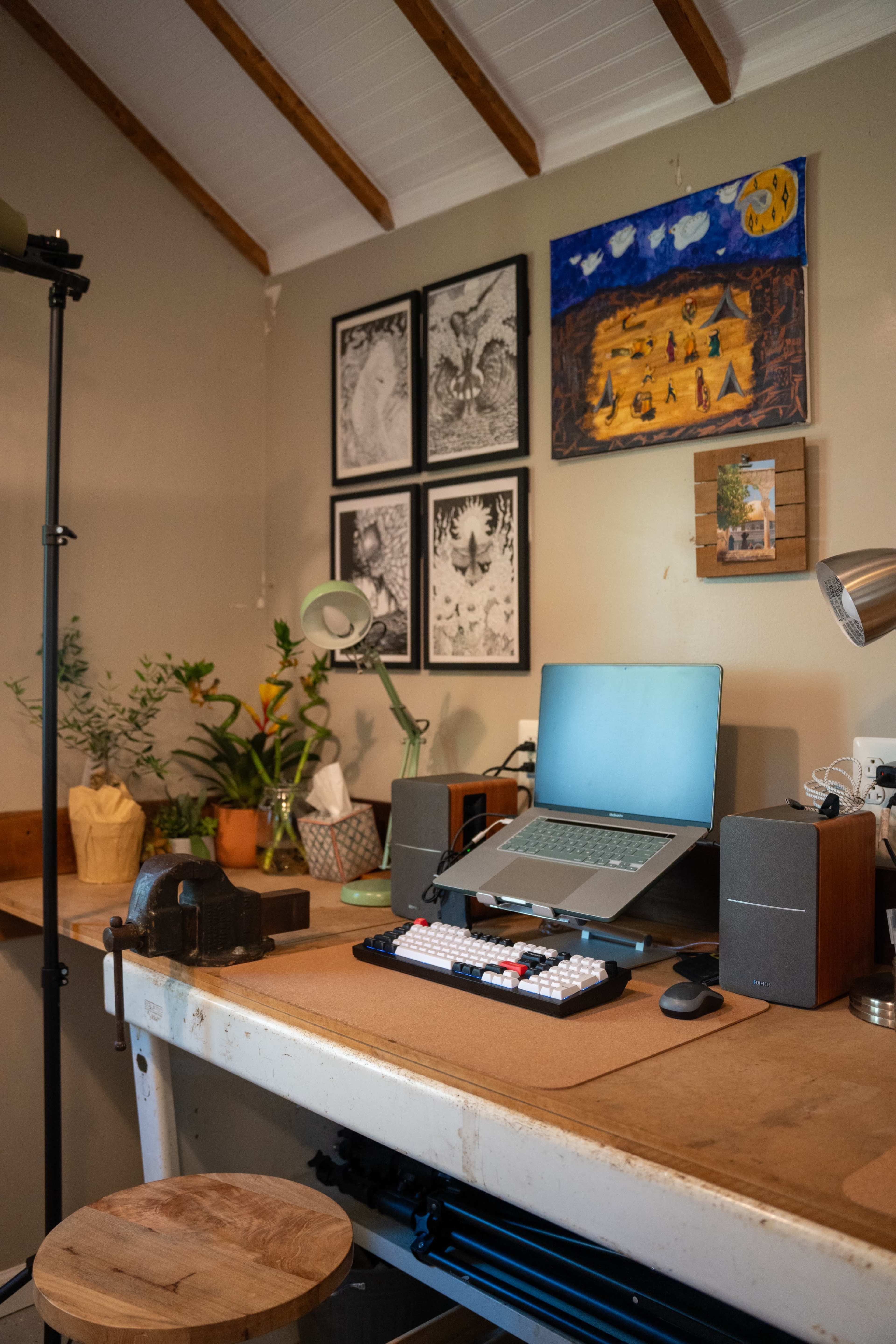 A wooden desk with a laptop, speakers, and various decorative items is set against a wall adorned with framed artwork and plants.