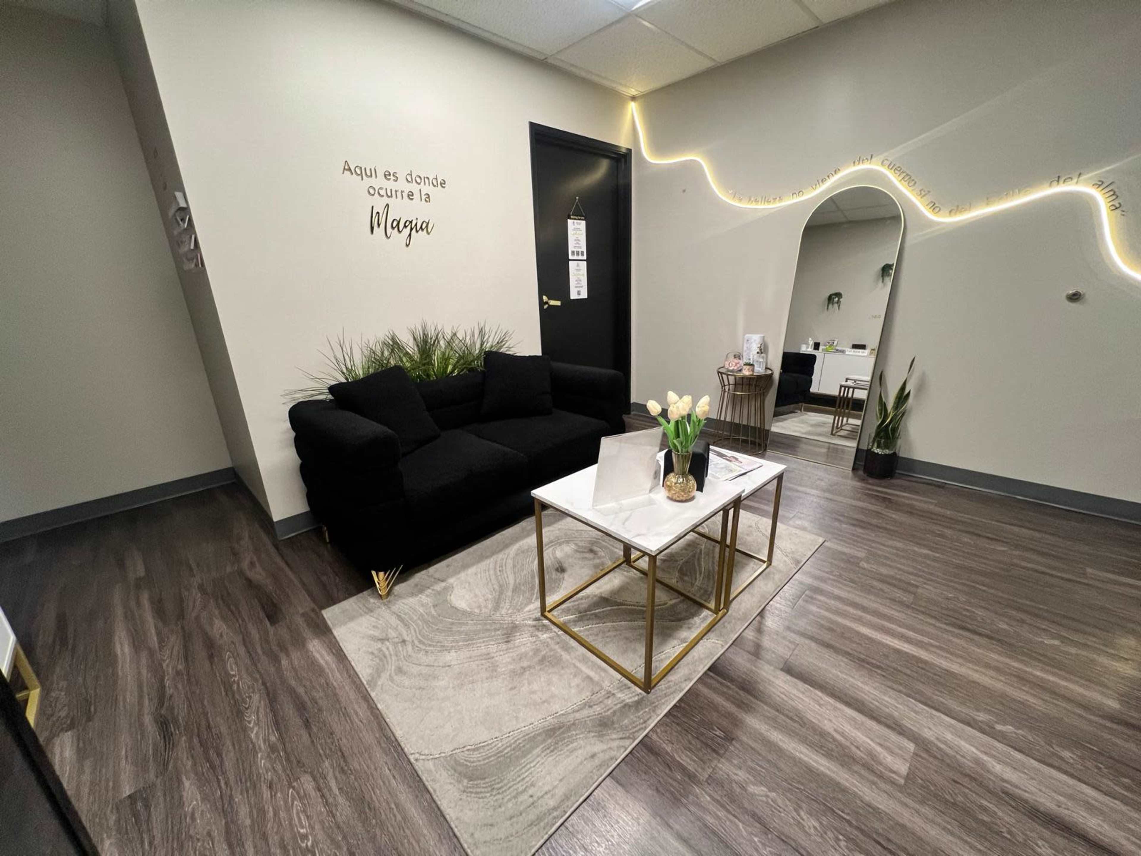 The image shows a modern waiting area with a black sofa, a coffee table, and decorative plants, featuring a wall with a Spanish phrase and soft lighting accents.