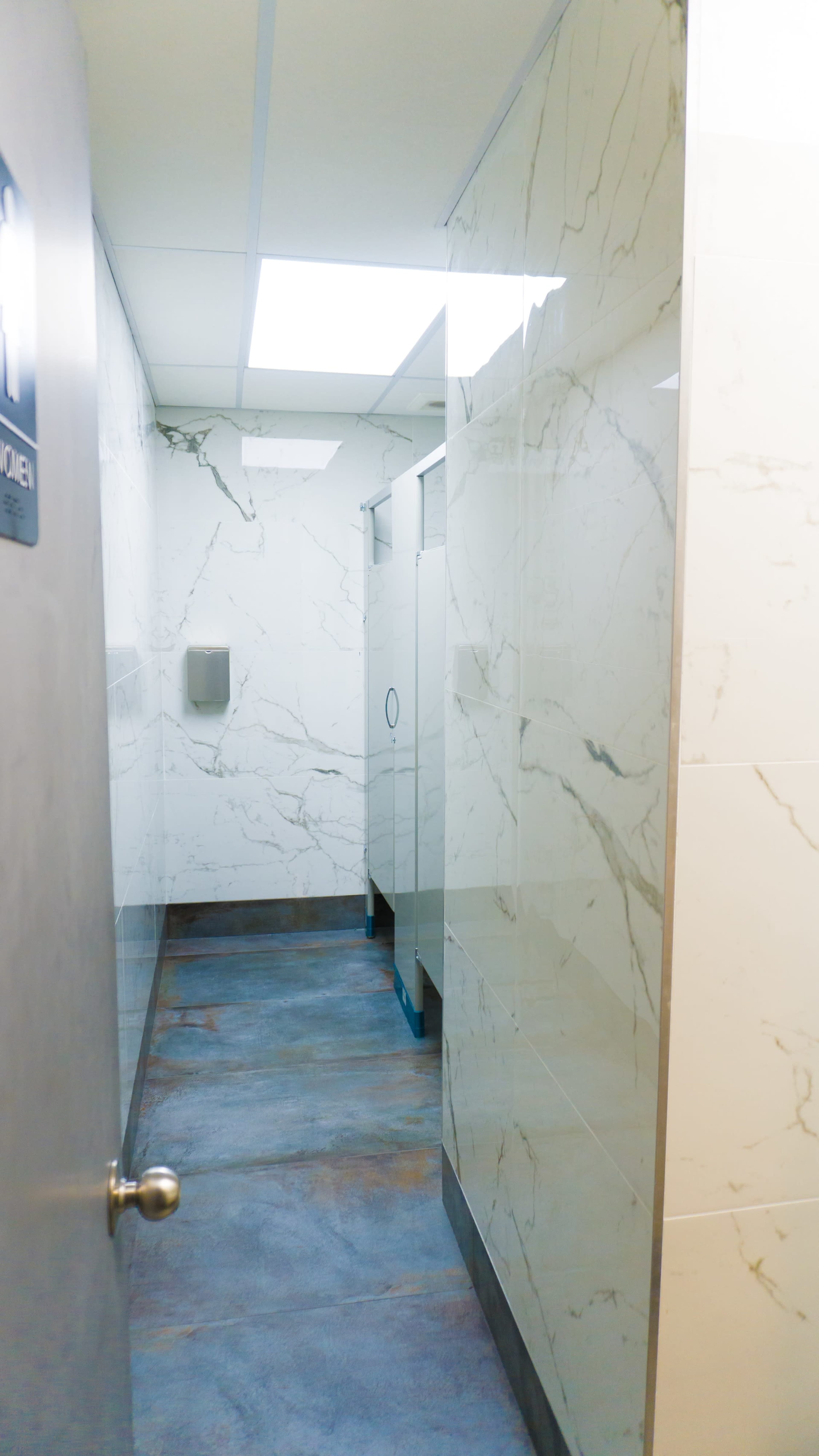 A well-lit restroom corridor features marble-patterned walls and stalls with a gray floor.