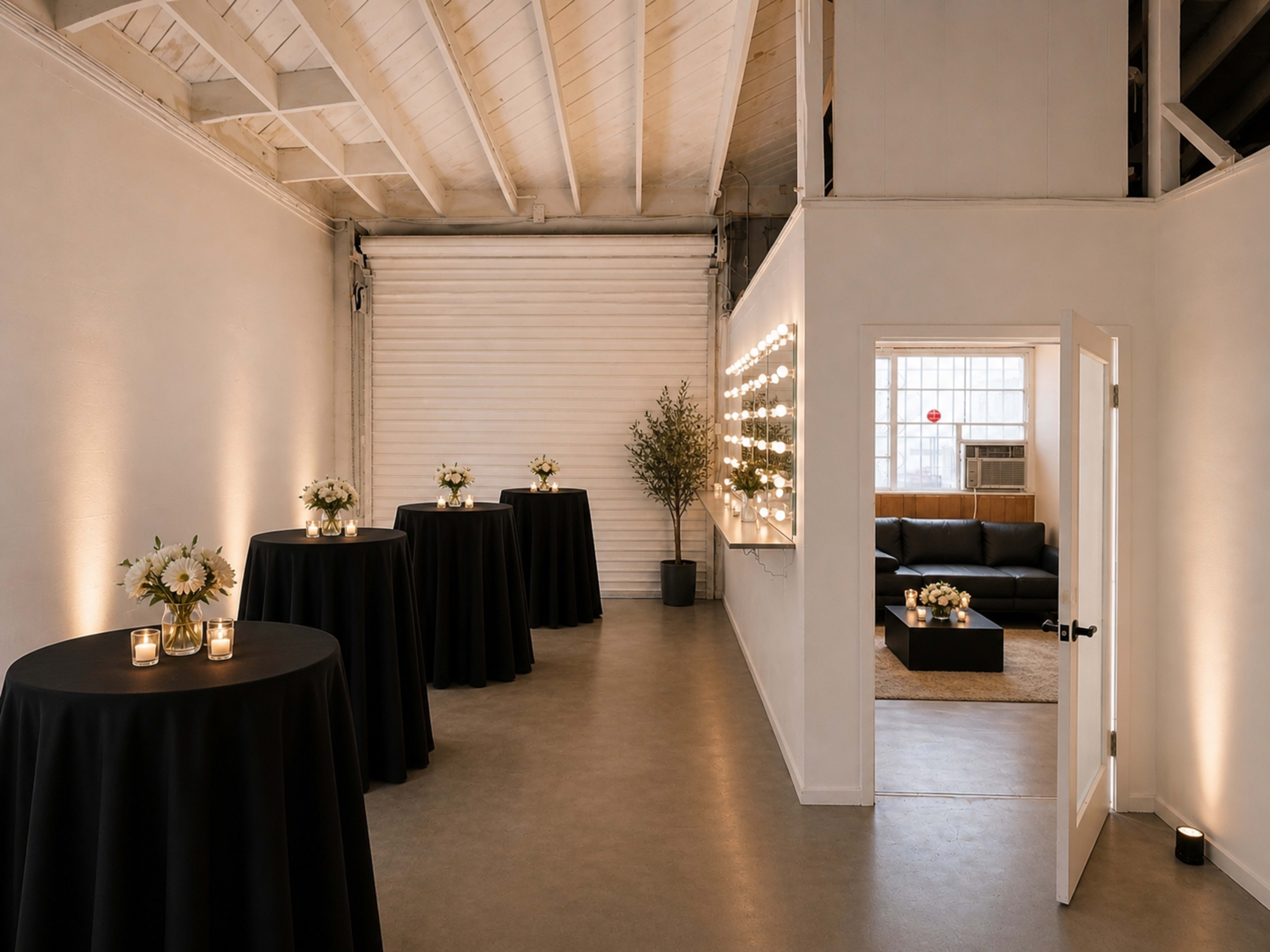 The image shows a modern, minimalist room with tall black tables adorned with floral arrangements and softly lit candles, leading to a cozy lounge area with a sofa in the background.
