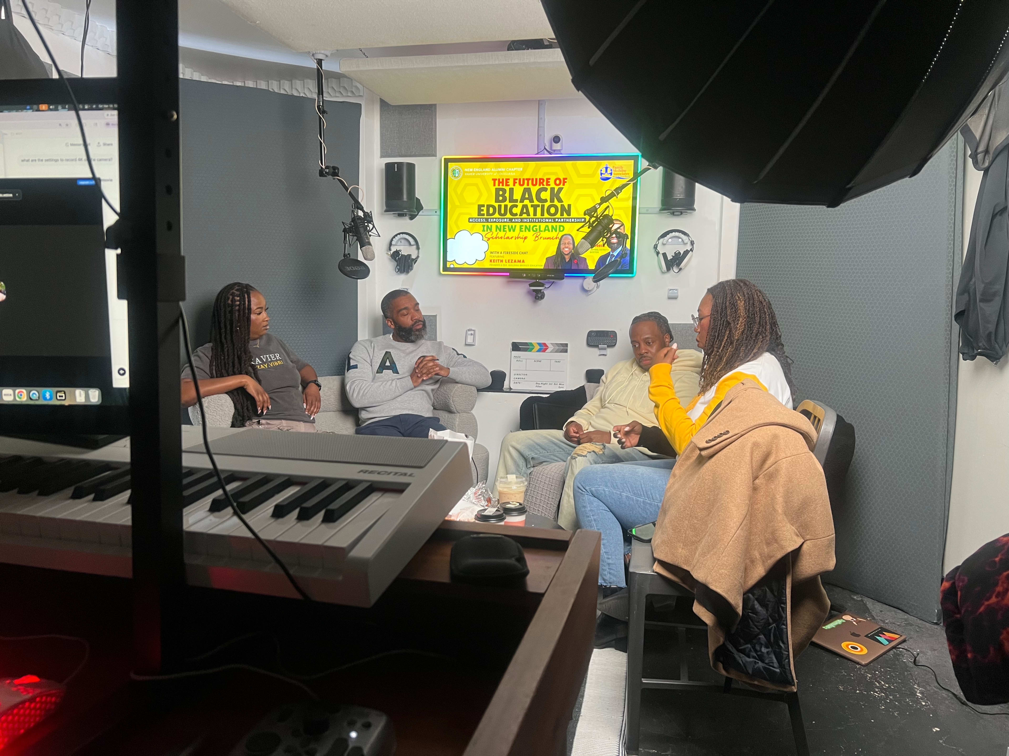 A group of four individuals engages in a conversation in a recording studio, with a colorful project screen displaying "The Future of Black Education" in the background.