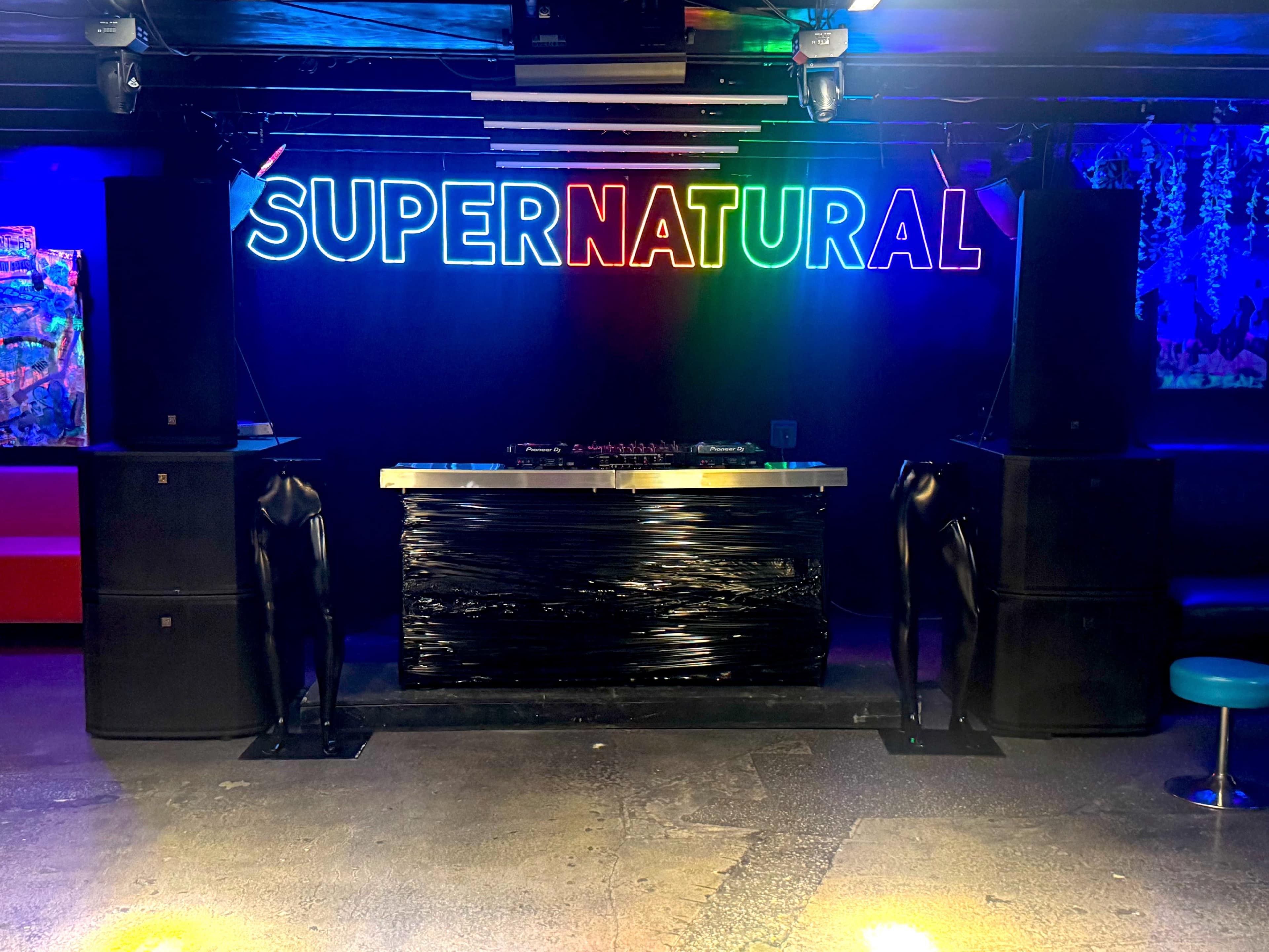 The photo shows a DJ booth with a neon sign that reads "SUPERNATURAL" and two black, humanoid sculptures flanking the booth.