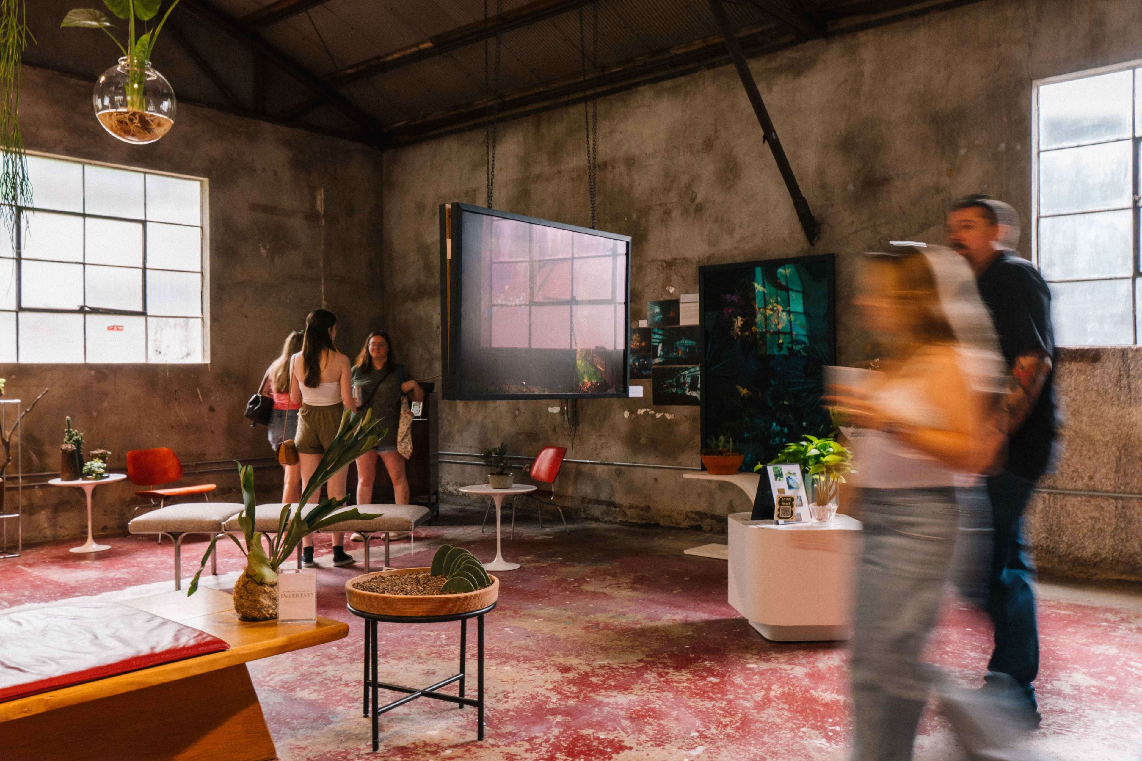 The scene shows a spacious, industrial-styled room with a mix of modern furniture, large windows, and people casually interacting while surrounded by potted plants.