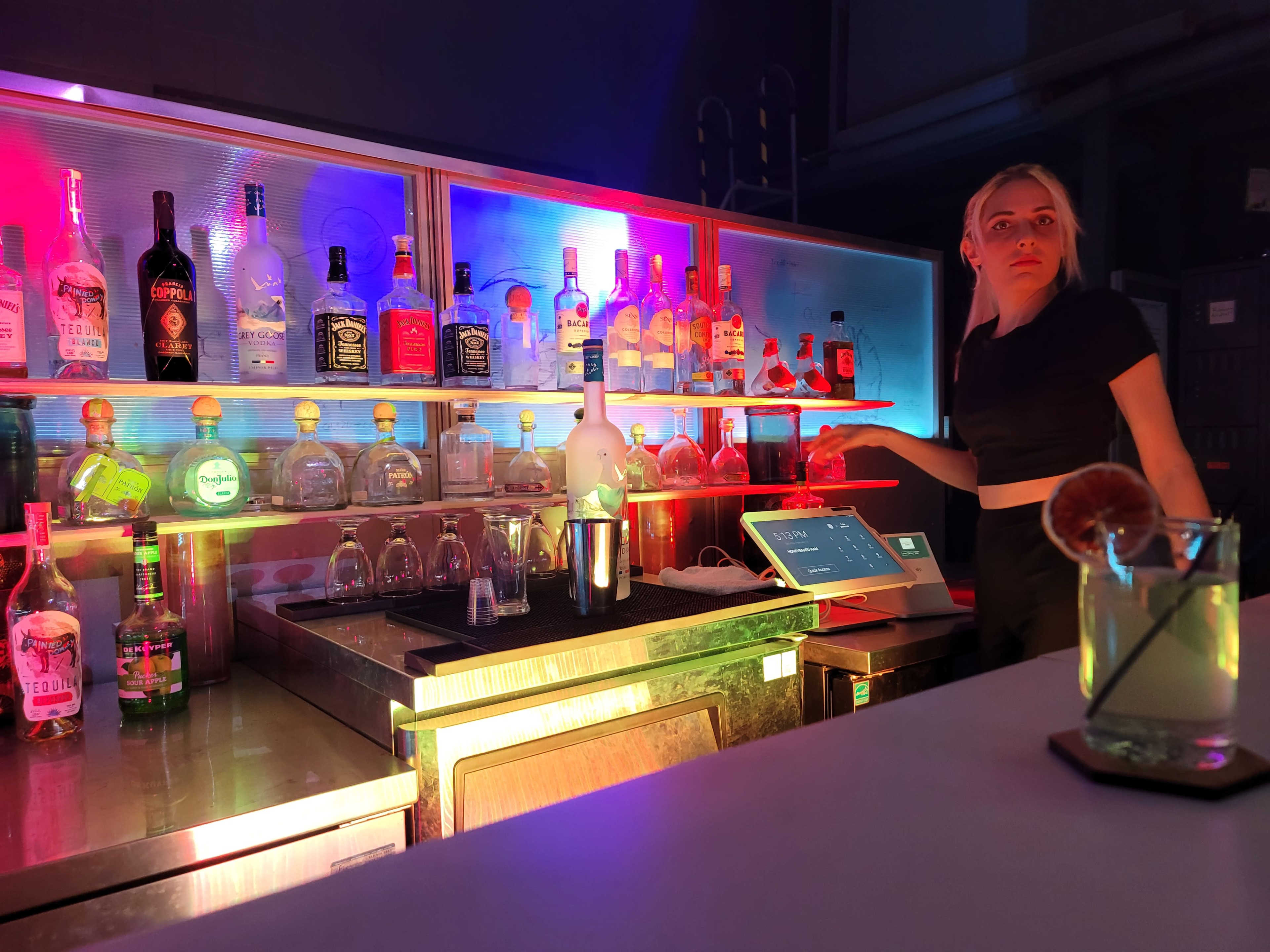 A bartender stands behind a brightly lit bar stocked with various bottles of liquor and colorful neon lights.