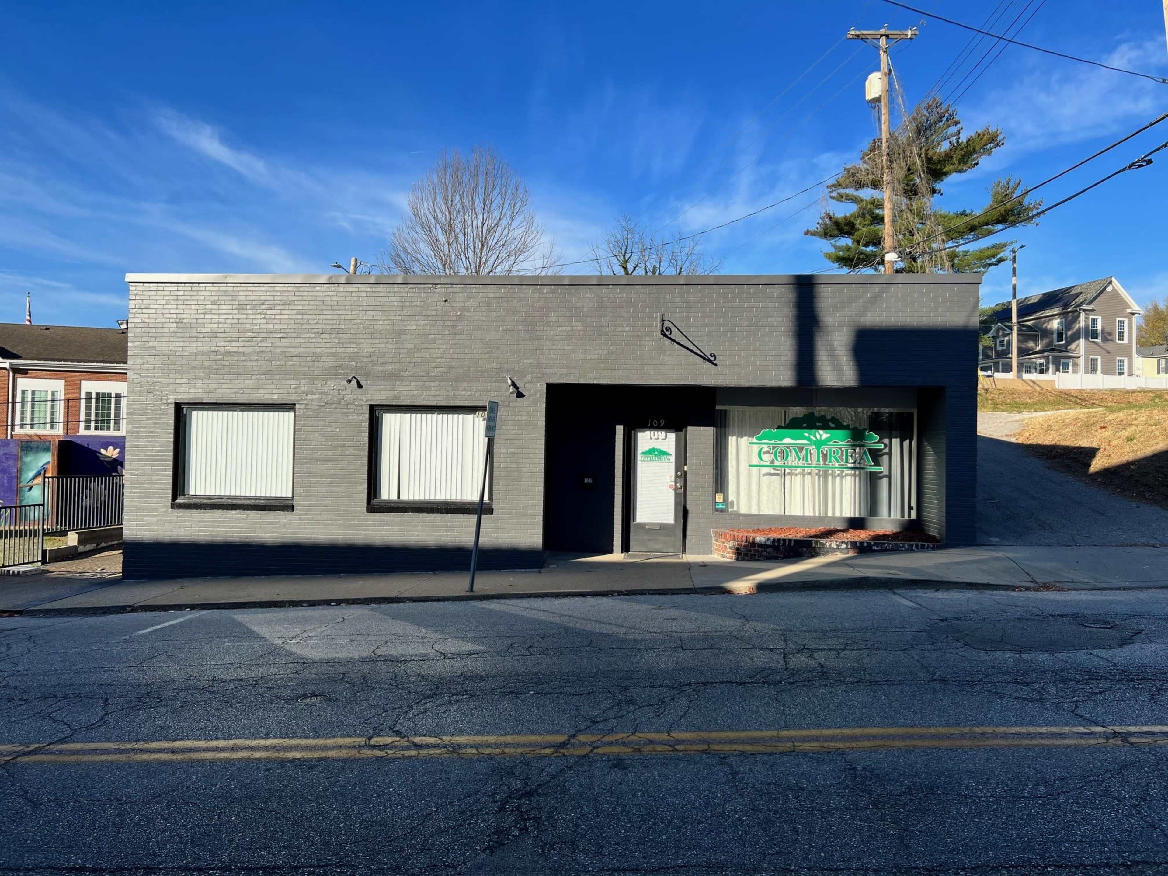 A single-story, black-brick building with large windows and a green sign, situated along a sloped street.