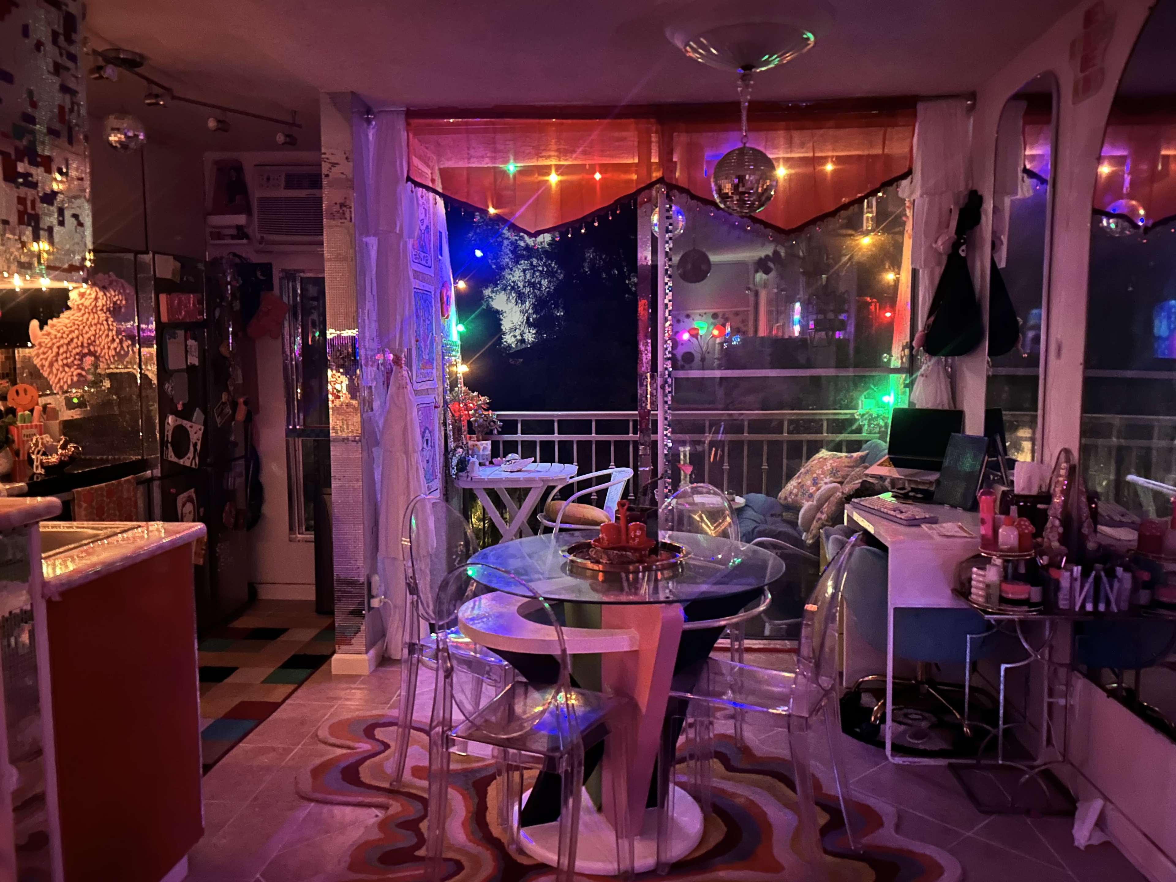 The image shows a brightly lit, eclectic living space with colorful decorative lights, a glass dining table surrounded by transparent chairs, and a cozy seating area.