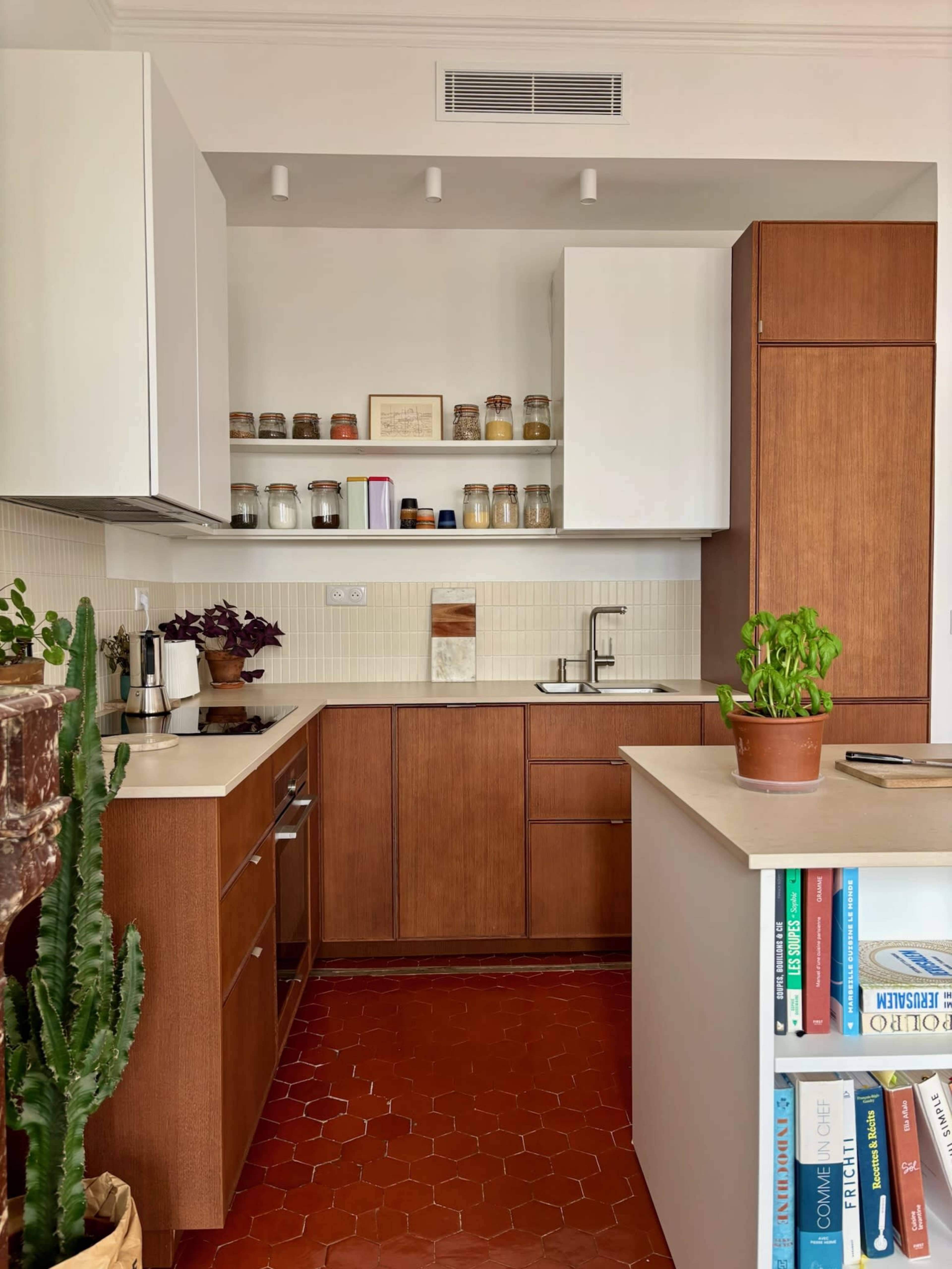 The image shows a modern kitchen with wooden cabinets, a white countertop, and a small table featuring a potted plant and books.