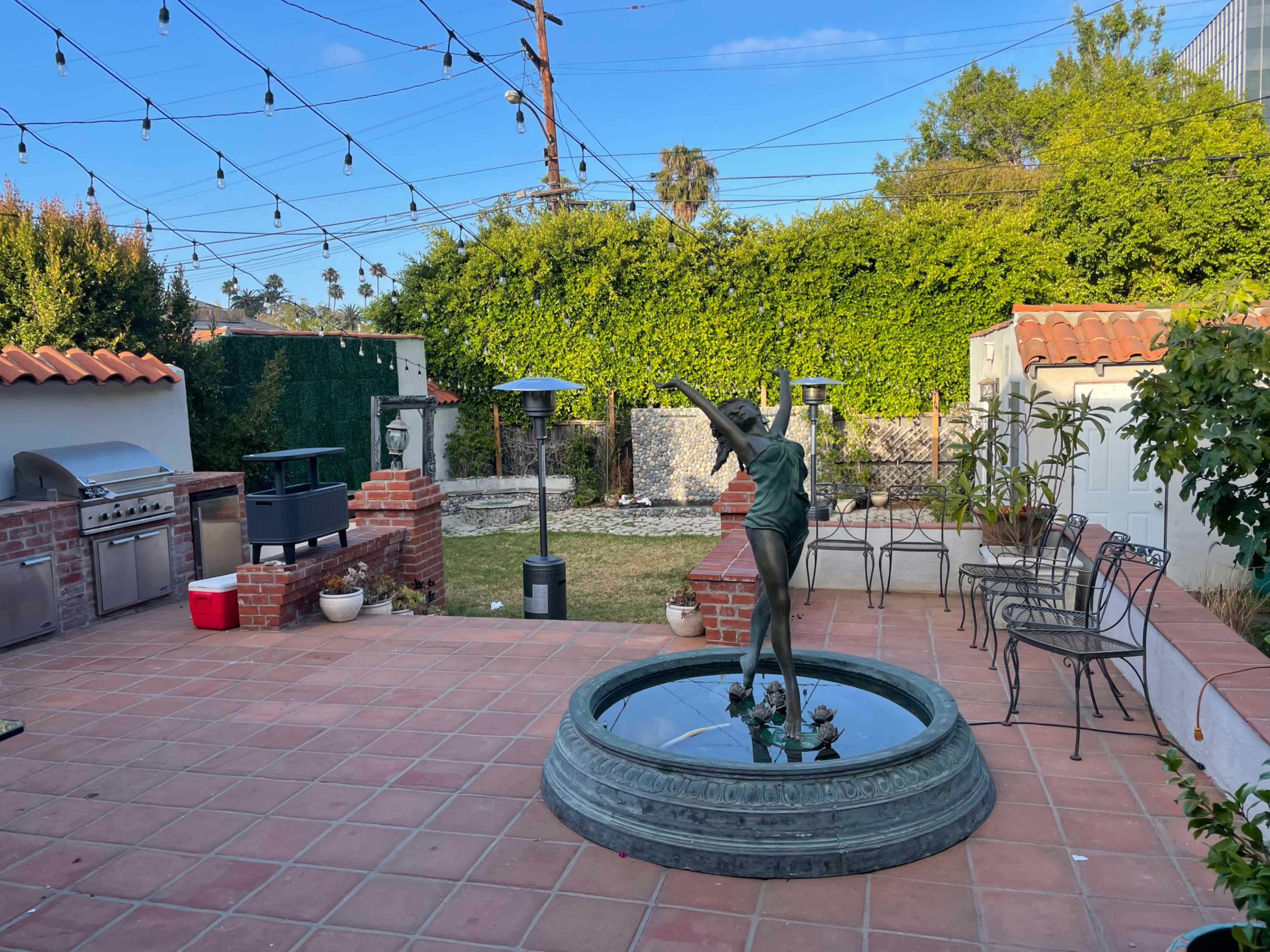 The image shows a backyard with a tiled patio featuring a statue in a fountain, surrounded by outdoor seating, a grill, and lush greenery under a clear sky.