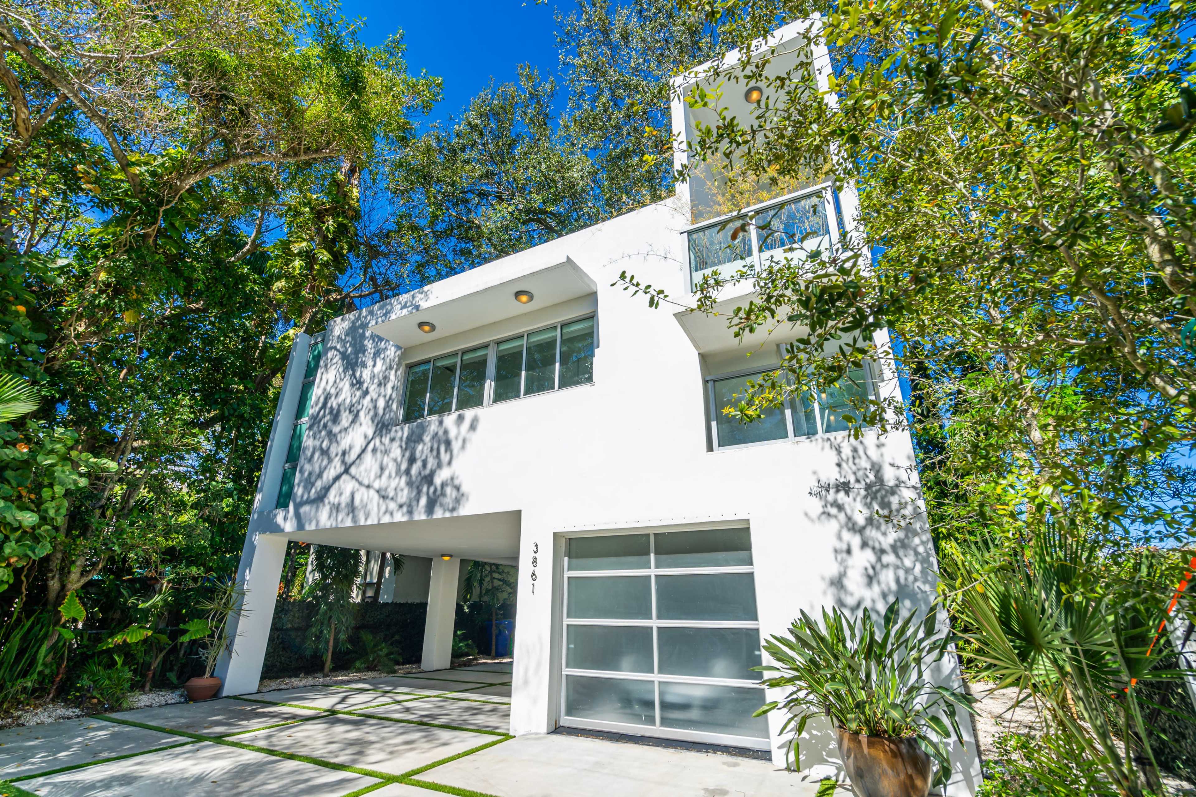A modern white three-story house surrounded by lush greenery and palm trees.