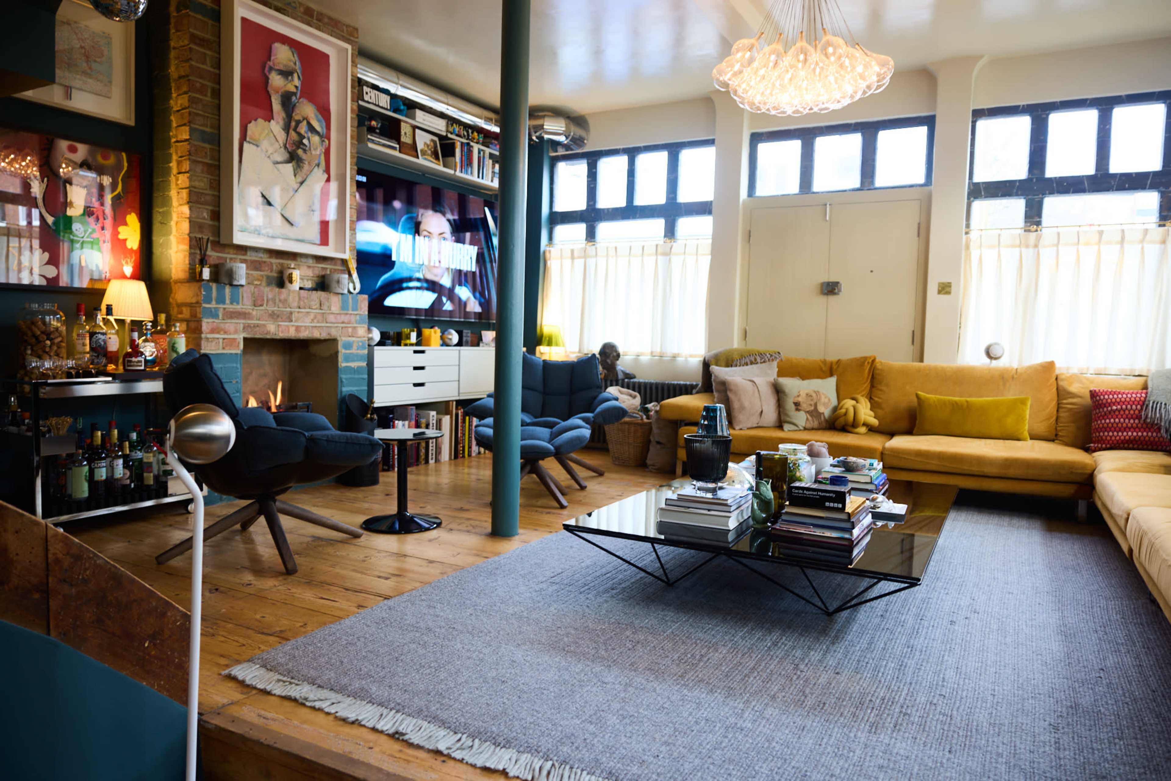 A modern living room with a mix of yellow and blue furniture, a fireplace, and large windows allowing natural light.