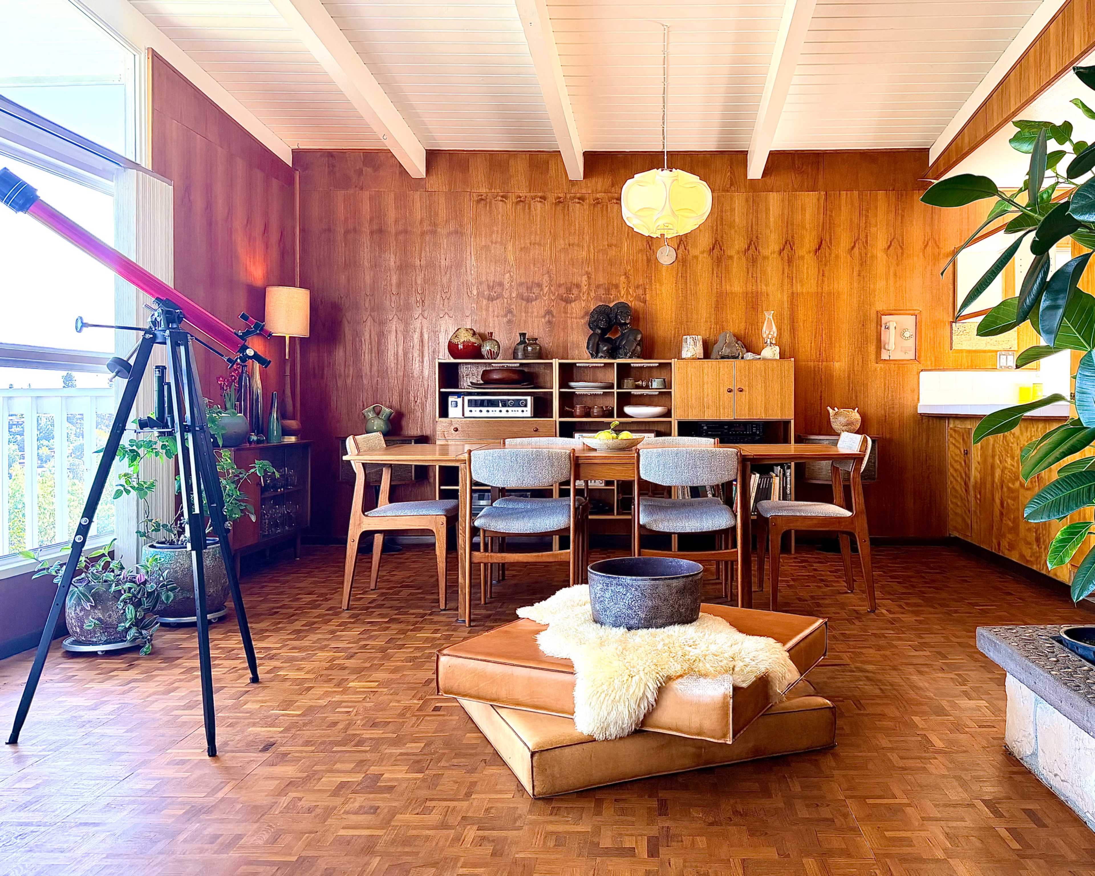 The image depicts a mid-century modern dining room featuring a wooden wall, a table with chairs, a telescope in the corner, and a cozy seating area with a round cushion and a planter.