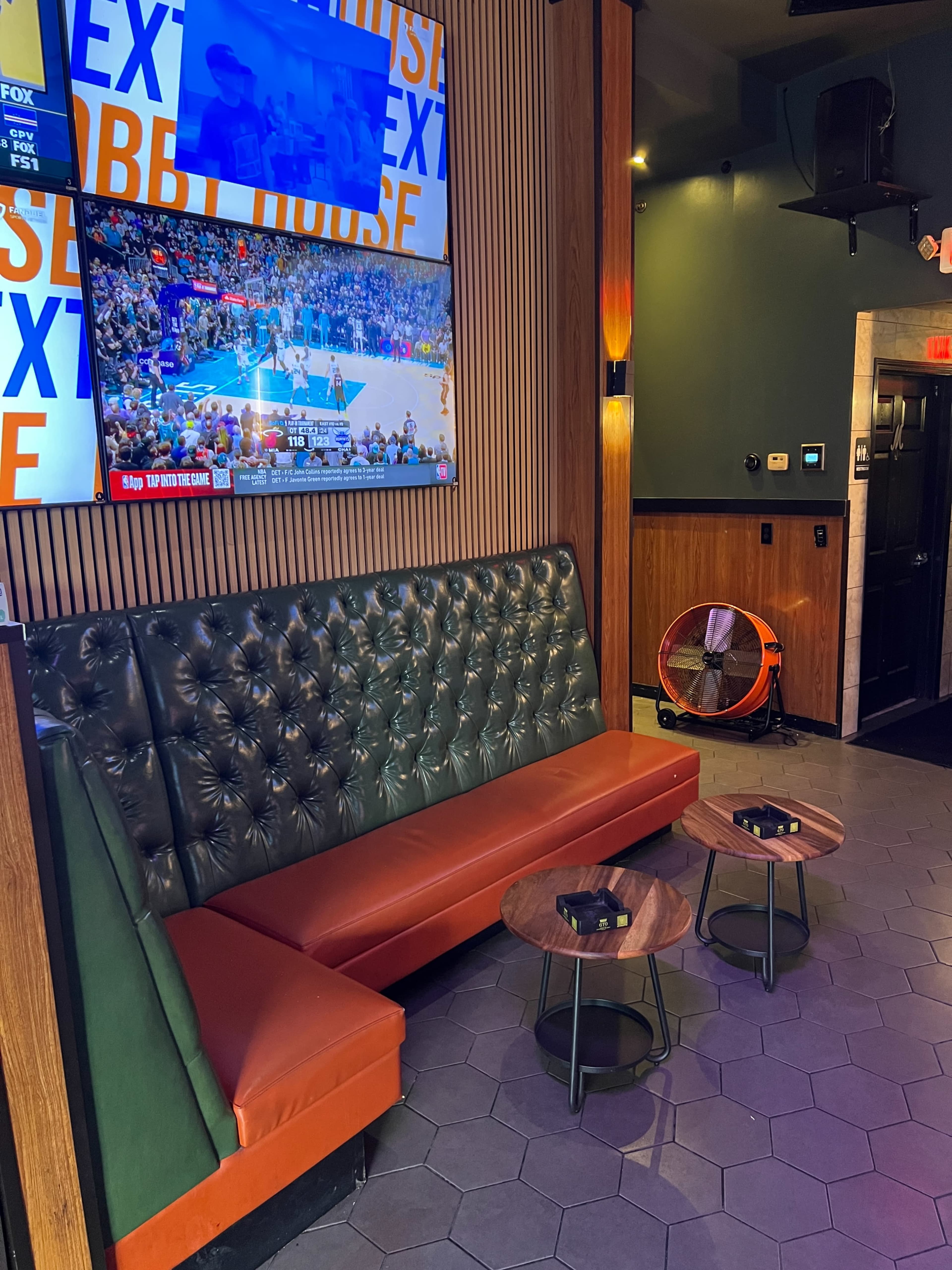 The image shows a cozy seating area in a bar, featuring a green tufted bench, two small wooden tables, and a television displaying a basketball game.